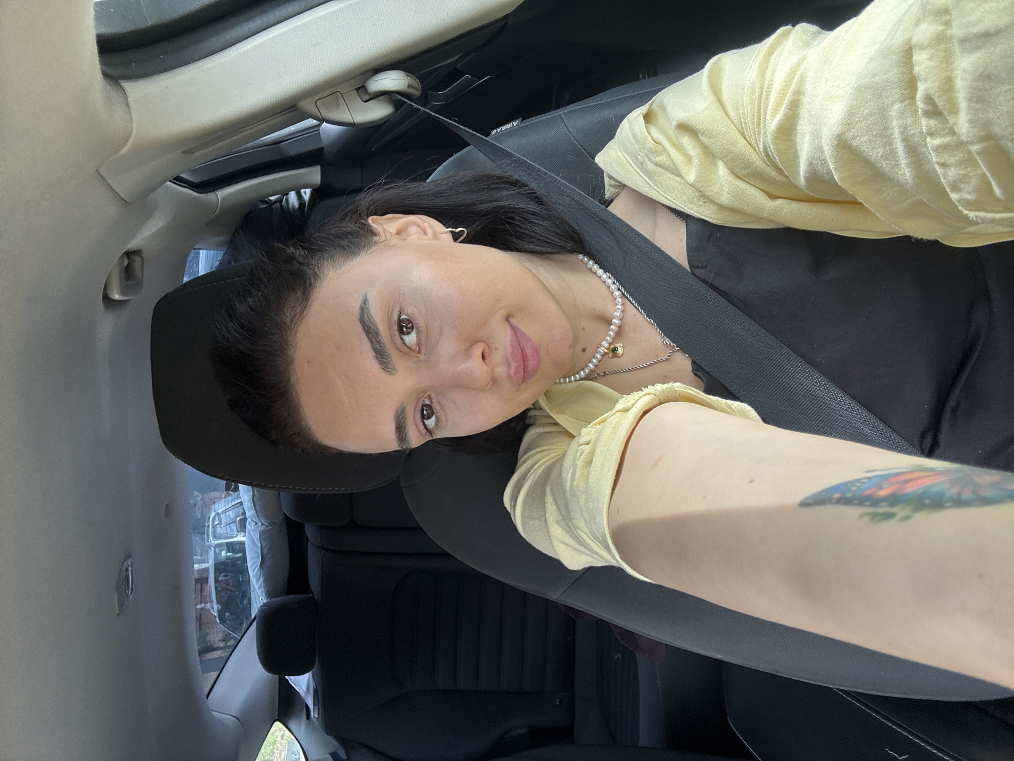 A woman is sitting in the driver's seat of a car, wearing a seatbelt. She is wearing a black top, a yellow cardigan, and two necklaces. The woman has dark hair and brown eyes. She has a butterfly tattoo on her left arm. The interior of the car is visible, including the dashboard and the back seat. The location outside the car is not identifiable as being in Tbilisi, Georgia based on the image provided.