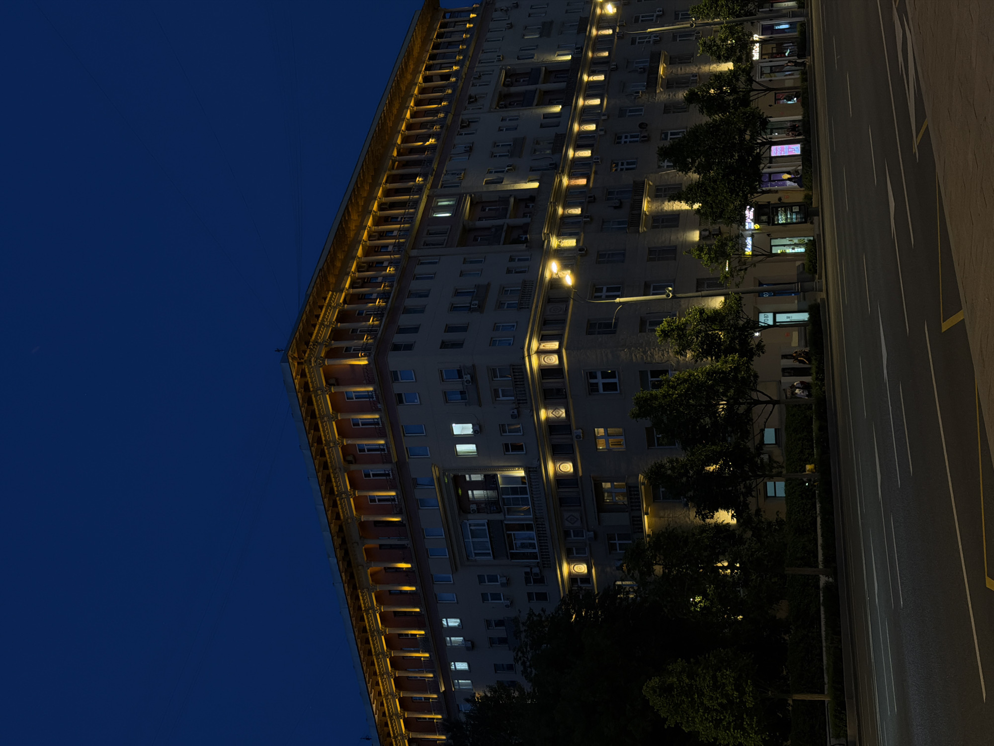 This night-time image captures a large, multi-story building, characteristic of Stalinist Empire architecture, located on a wide urban street in Moscow, Russia. The building features an illuminated grand colonnade along its upper levels, with warm yellow lights highlighting the architectural details. Below, numerous windows display varying levels of interior light, some appearing white or cool blue. The building's façade is adorned with additional decorative elements and balconies, many with visible air conditioning units.
The ground floor hosts several commercial establishments, identified by their brightly lit storefronts. One visible sign in Cyrillic reads "Скупка Золота" (Gold Purchase), and another appears to be for "ВТБ" (VTB bank).
In the foreground, a multi-lane asphalt road with clear white lane markings and a yellow median line stretches across the frame. A sidewalk runs parallel to the building, populated by evenly spaced, leafy deciduous trees. Several streetlights illuminate the road and pedestrian areas. A few indistinct human figures are visible on the sidewalk near the commercial units. The sky is a deep, dark blue, indicating the late hours of the night.
The architectural style and commercial signs strongly suggest the location is a prominent avenue in Moscow, specifically the building at Tverskaya Street 15, known for its distinctive Stalinist neoclassical design and illuminated colonnade.