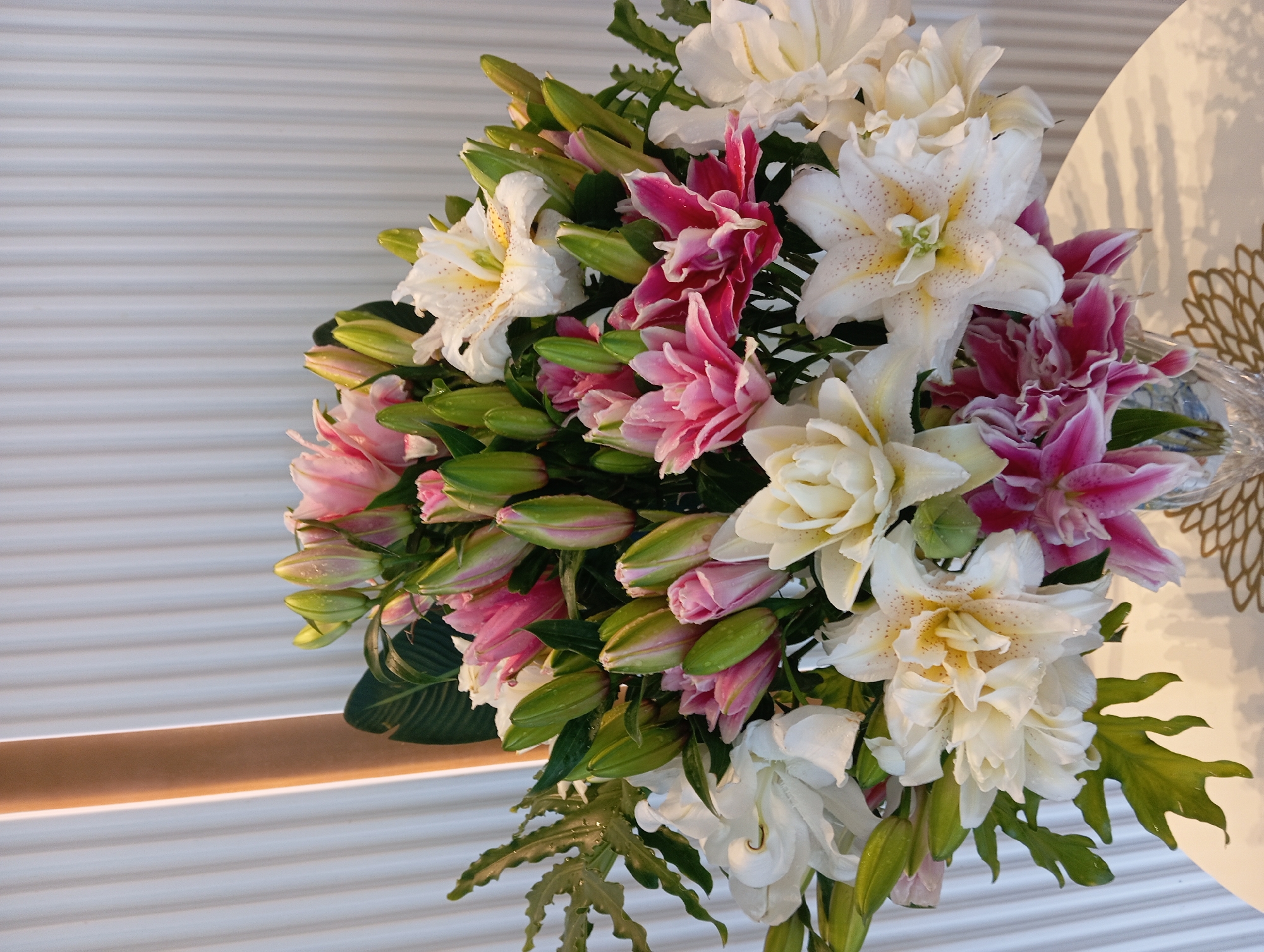A close-up, eye-level shot features a large bouquet of lilies in various stages of bloom, predominantly white and pink. The lilies are double-petaled and some have speckled stamen. They are arranged in a clear glass vase, which rests on a white table with a textured surface. The background consists of vertical, pale gray blinds illuminated by a soft, warm light source to the left, suggesting an indoor setting, possibly a hotel room or a private residence in Nha Trang, Vietnam. There are no people or animals present in the image.