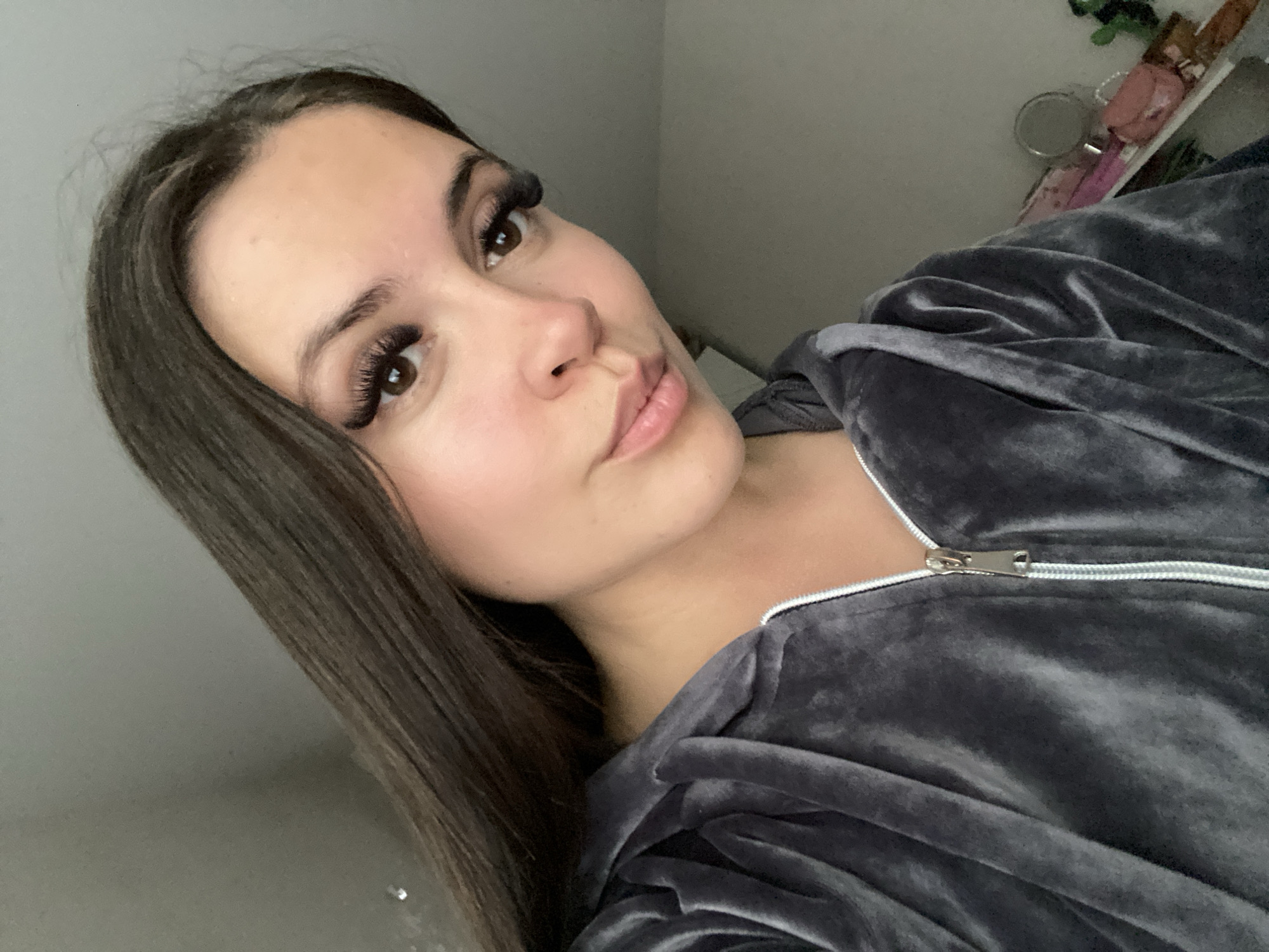A young woman is depicted in a close-up, angled selfie, taken indoors. She has fair skin, long, straight dark brown hair that falls over her shoulders, and brown eyes. Her gaze is directed towards the camera, and her lips are slightly parted, exhibiting a light pink color. She has prominent, voluminous eyelashes, which appear to be extensions.
She is wearing a dark grey, soft-textured garment, possibly velvet or velour, which appears to be a hooded top or jacket. A silver zipper on the front of her clothing is partially unzipped. The image is framed from a slightly high and tilted angle, focusing on her face and upper body.
The background is softly blurred but indicates an indoor setting, likely a room. A light-colored wall is visible behind her, and to the right, indistinct objects are discernible on what appears to be a shelf or surface. These include a small pink item, possibly a bag or decorative piece, and a circular reflective object, perhaps a small mirror, along with a hint of green. The lighting is soft and even, highlighting her features.
The woman is posing for the photograph with a calm and relaxed expression. No text is visible in the image. The location context for this scene is Chaikovsky, Russia.