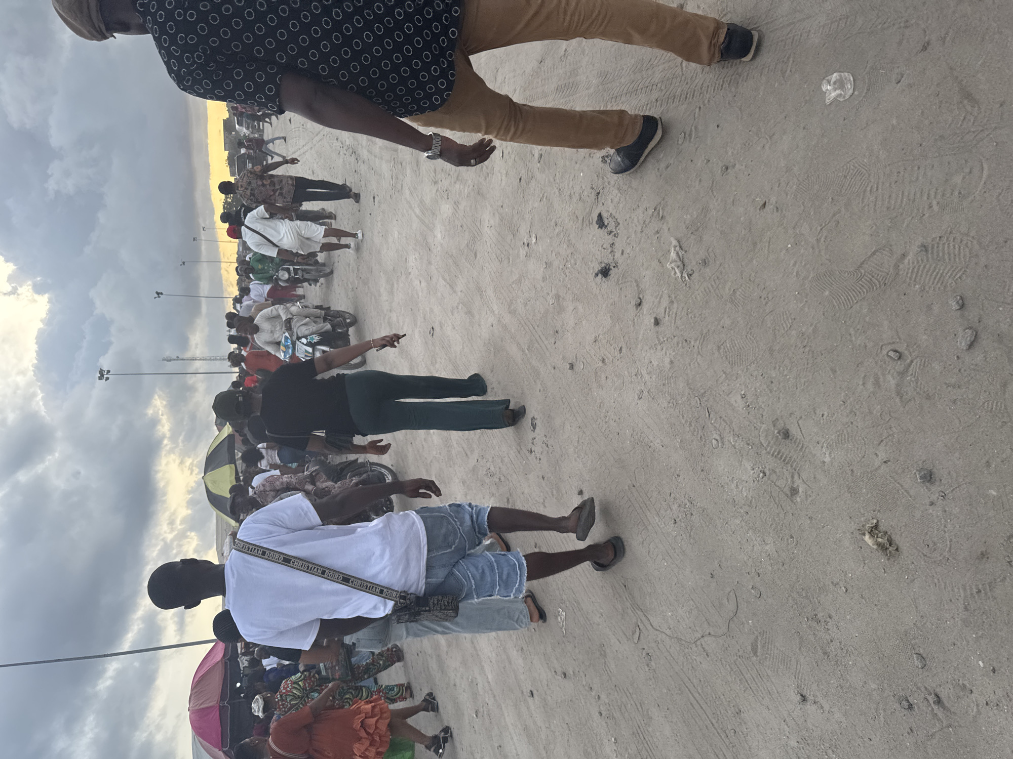 The image depicts a gathering of people on a sandy, outdoor area, likely in Satellite Town, Nigeria. The environment consists of a wide-open space with an overcast sky. Several people are visible, with one person in the foreground wearing a white shirt, denim shorts, and sandals. Another person in the upper frame is wearing a patterned shirt and brown pants. Several other people are in the background, with some appearing to be on motorcycles. There are streetlights visible. The sandy ground shows tire tracks and scattered debris.