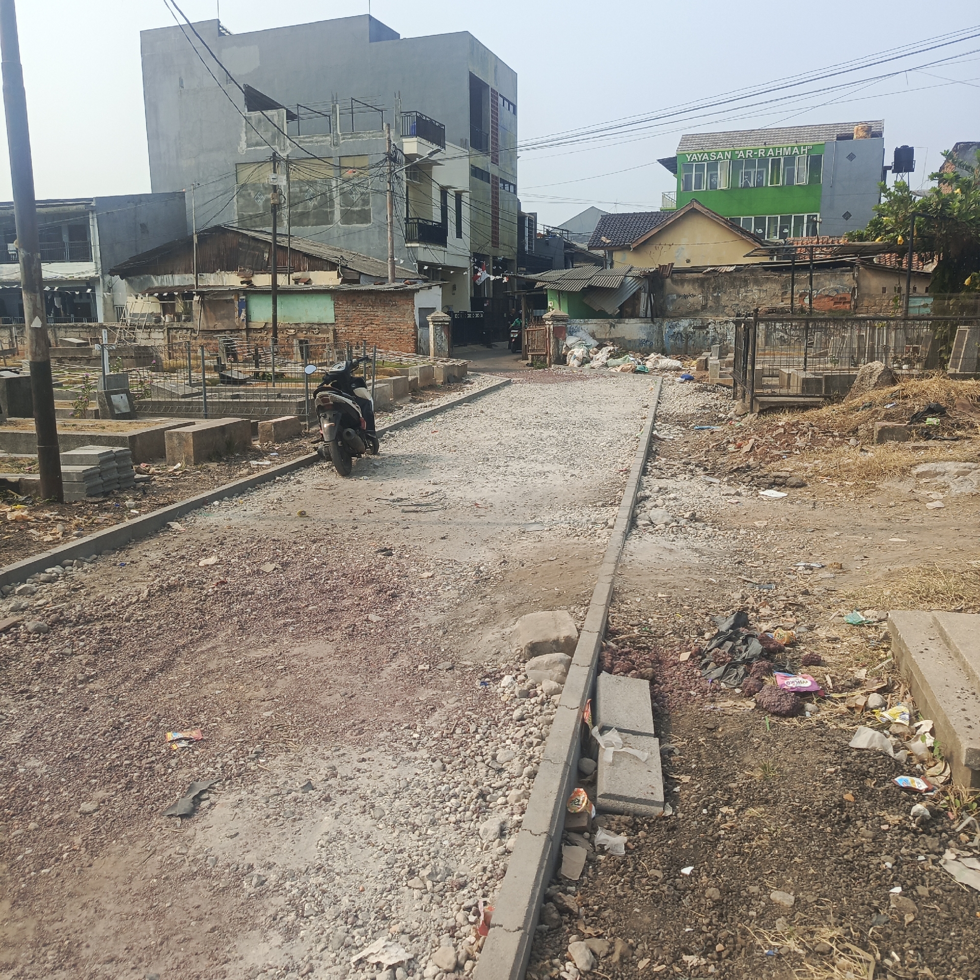The image captures a scene of ongoing road construction in a densely populated urban area. A gravel-covered road is under construction, with visible building materials such as concrete blocks and loose gravel. A black scooter is parked on the side of the road. In the background, a multi-story building with a green facade and signage reading "YAYASAN 'AR-RAHMAH'" suggests the presence of an educational or community institution. Surrounding buildings appear aged and varied in architectural style. The environment is characterized by a mix of construction debris and accumulated litter, indicating a somewhat unkempt public space. The overall impression is one of urban development and routine life coexisting with the disruption of construction.