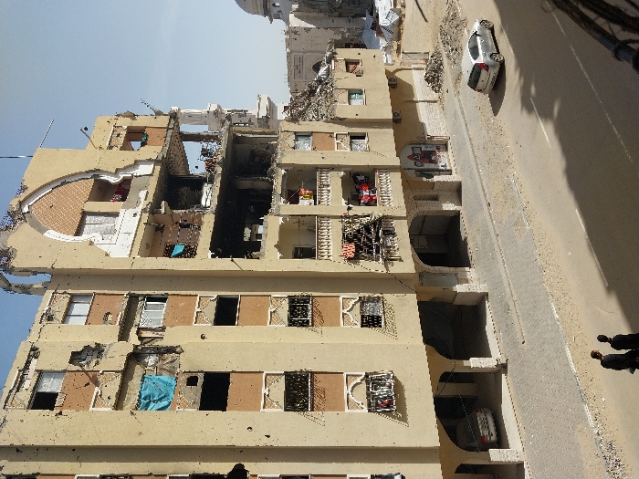 This image presents a severely damaged multi-story building located in Khan Yunis, Palestinian Territory, under a clear blue sky. The structure, appearing to be residential or mixed-use, features a light-tan facade with brown accent panels and exhibits extensive structural compromise. Large sections of its exterior walls are absent, exposing internal floors and rooms containing debris. Numerous windows are shattered or missing, and the building's upper right section shows a large, broken arched architectural feature, revealing internal construction. The facade is marked by multiple impact points consistent with bullet holes or shrapnel. Laundry hangs from several balconies, and a blue tarp is draped from an upper-floor window. The ground floor incorporates arched bays, one housing a white sedan. Adjacent to the building is a paved sidewalk, followed by a street with a sandy median or shoulder. In the foreground, two individuals dressed in dark attire walk away from the observer on the paved street. A silver hatchback is parked on the sandy shoulder to the right. Piles of construction debris are visible at the building's base on the right and further into the background, where a white domed structure with a minaret, possibly a mosque, is also visible amidst rubble.