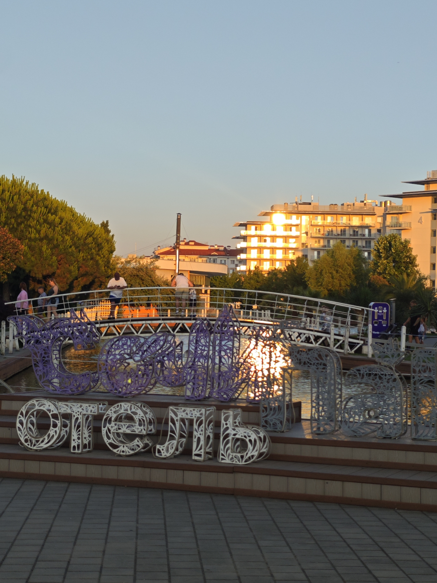 In Sochi, Russia, at sunset, a bridge spans a body of water. Several people are walking on the bridge. In the foreground, large decorative letters spelling out "ОТЕЛЬ" (Hotel) are displayed. Behind the bridge and water, there are trees and modern buildings, one of which is illuminated by the setting sun. The sky is clear and blue.