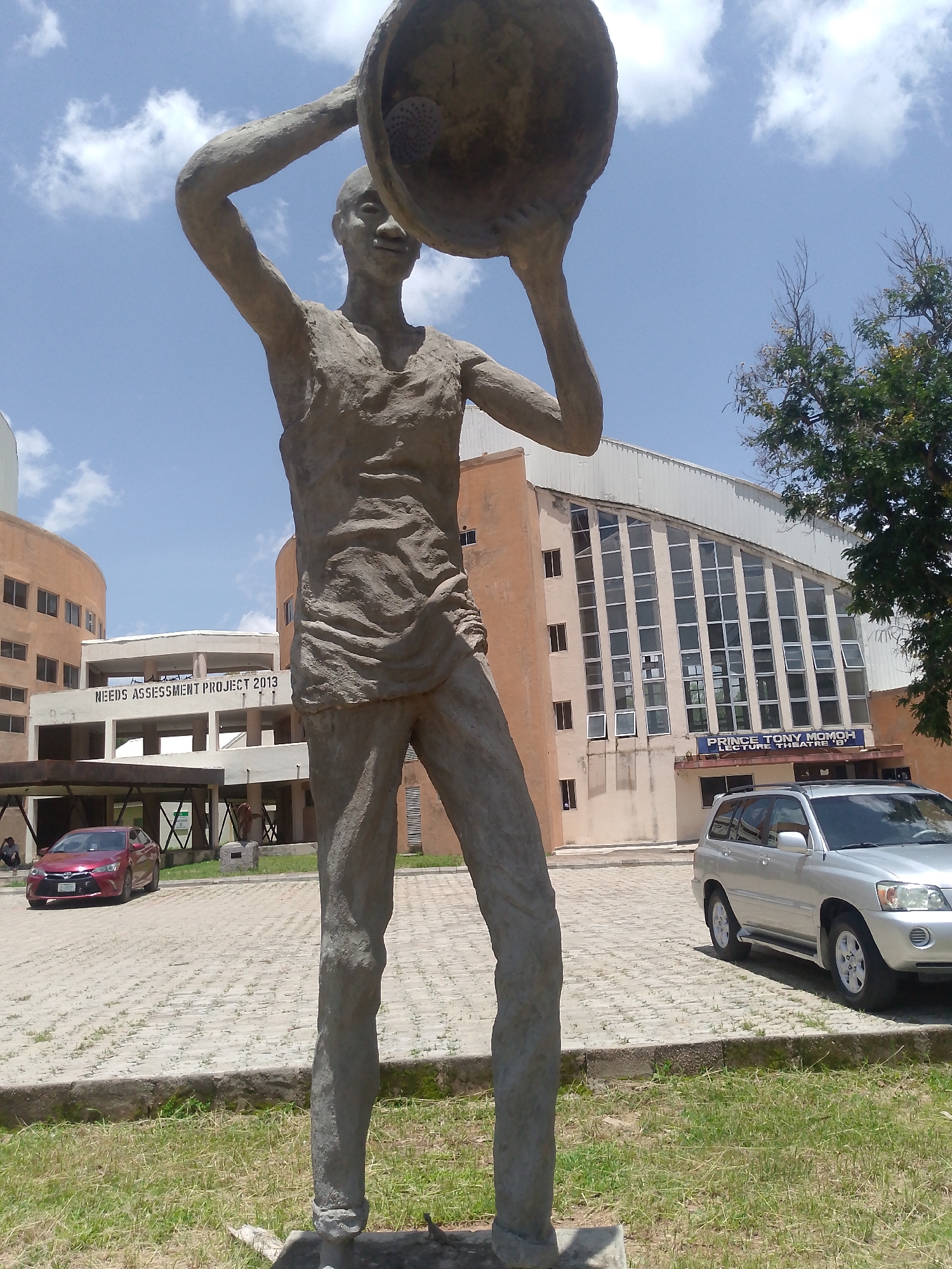 This image depicts a stone statue of a person holding a large, circular object above their head in an outdoor setting. The statue is in the foreground, with a paved courtyard and several buildings in the background. The buildings appear to be part of an institutional complex. One building has a sign that reads "PRINCE TONY MOMOH LECTURE THEATRE 'B'". Another building has a sign that reads "NEEDS ASSESSMENT PROJECT 2013". A red car is parked on the left side of the image, and a silver SUV is parked on the right. The sky is blue with some clouds. The location is identified as Barakin Angundi, Nigeria.