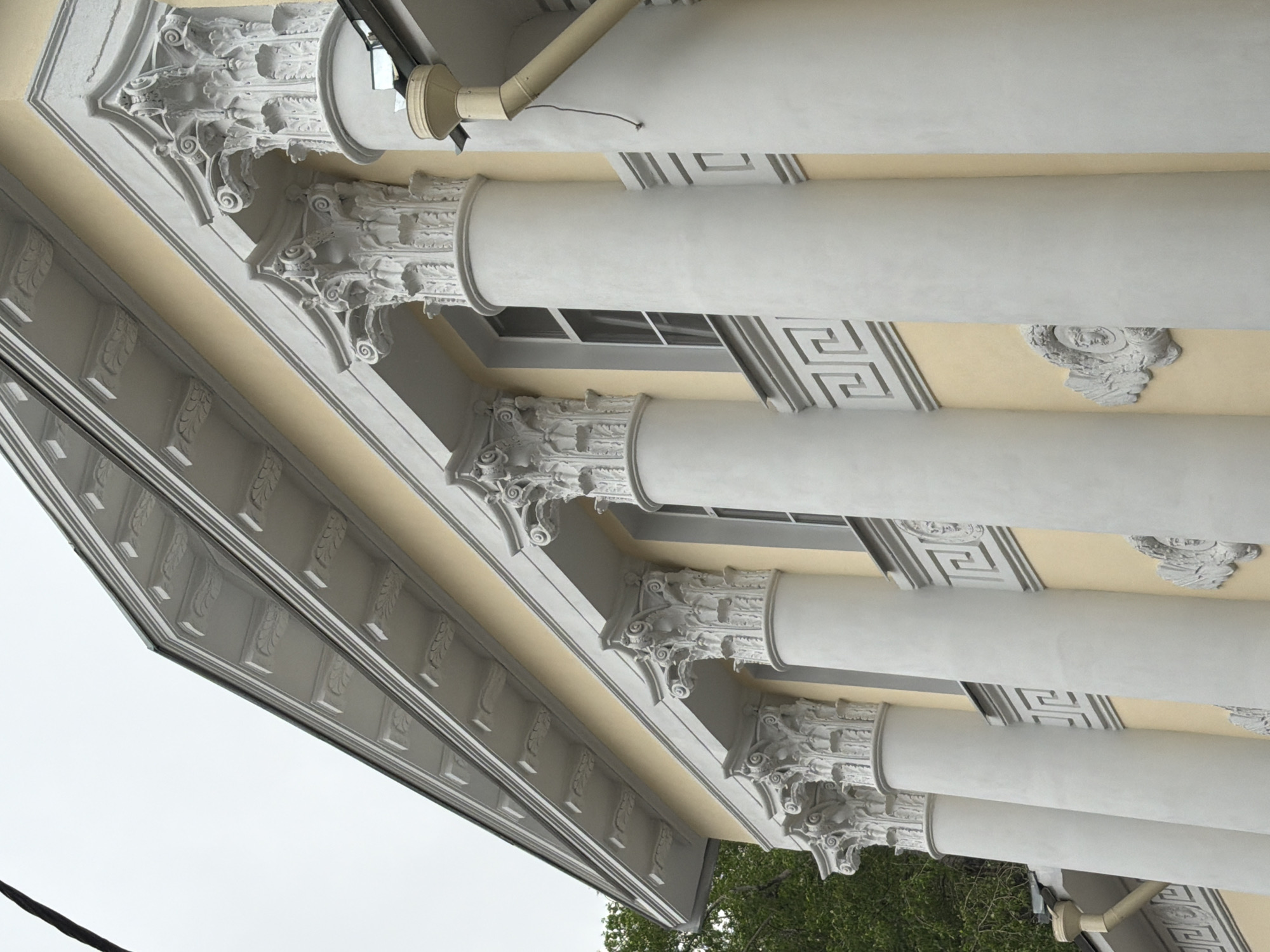 The image shows a portion of a building's exterior, specifically the columns and decorative elements of what appears to be a classical or neoclassical architectural style. The columns are white and cylindrical, with ornate capitals. The building's facade is a light yellow color. The elements have Greek key patterns and relief sculptures of faces. A drain pipe is visible near the top of the frame. The sky is overcast, suggesting a cloudy day. The bottom of the frame shows some greenery from nearby trees. I am unable to identify the precise location in Moscow.