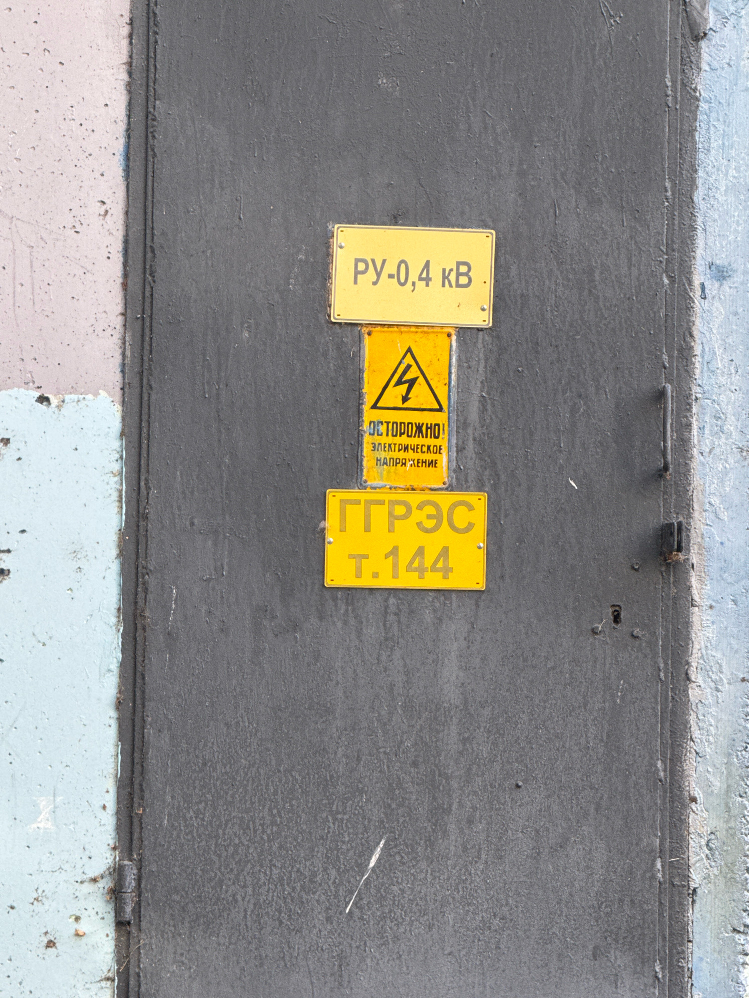 A dark gray metal door is mounted on a concrete wall. Three yellow signs are affixed to the door. The top sign reads "РУ-0,4 кВ". Below it, a triangular sign with a lightning bolt symbol and the words "ОСТОРОЖНО! ЭЛЕКТРИЧЕСКОЕ НАПРЯЖЕНИЕ" (Danger! High Voltage) is mounted. The bottom sign displays "ГРЭС Т.144". The door has a handle and a lock. The wall to the left of the door is painted in a light gray and blue mottled pattern. The wall to the right is rough concrete. The scene appears to be an exterior view of an electrical equipment enclosure or utility building.