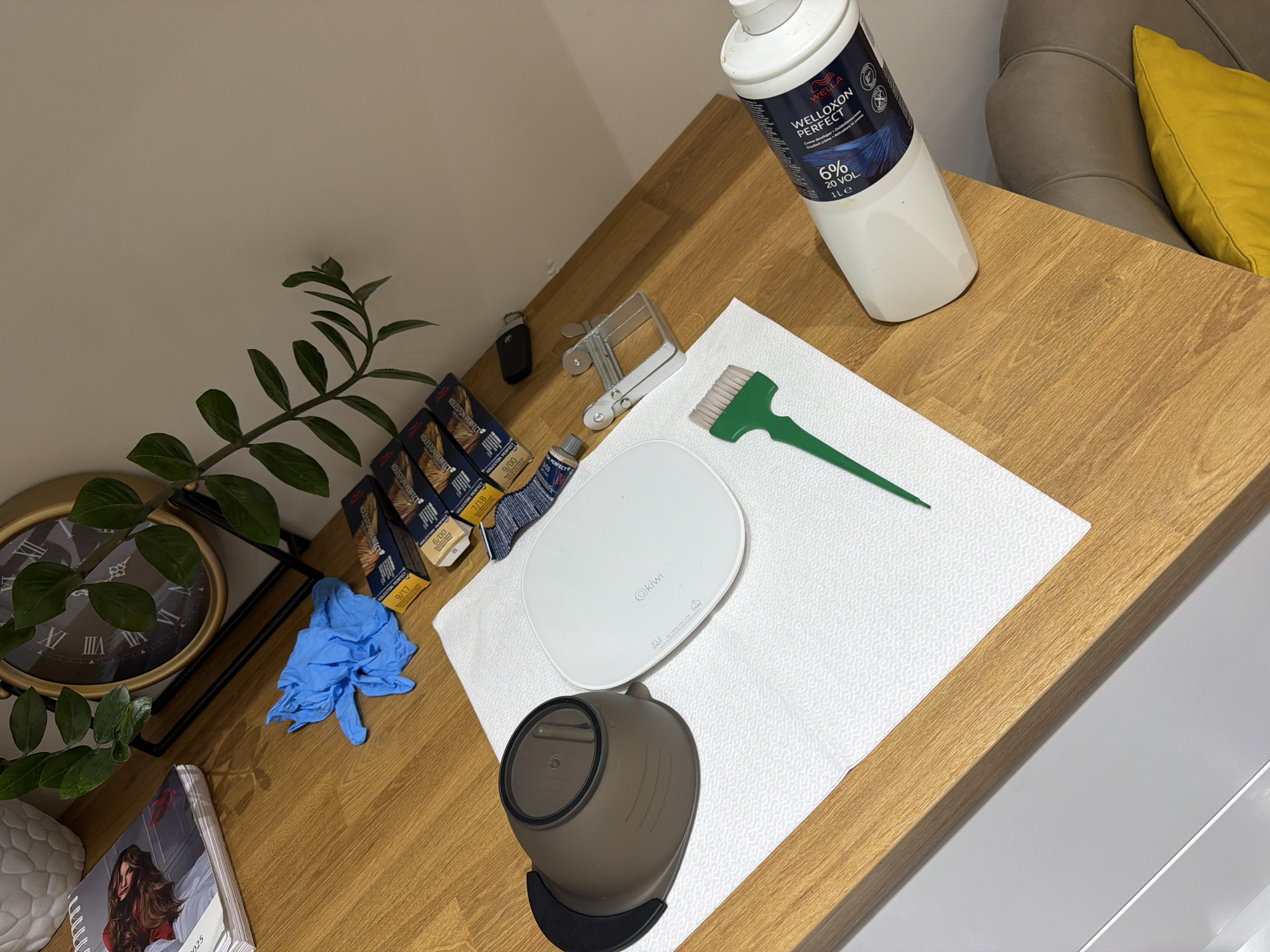 The image depicts a wooden countertop with various items related to hair dyeing. In the upper left, a white textured pot and a stack of papers with an image of a woman's hair are visible, along with a gold-framed clock in the background. Several boxes of hair dye, identified by the brand "Koleston Perfect," are arranged on the counter. A pair of blue nitrile gloves is also present.
In the center of the image, a brown mixing bowl and a white scale are placed on a folded white paper towel. Next to these is a green-tipped hair dye brush. To the right, a clear plastic clip dispenser and a small black object, possibly a key fob, are situated. Dominating the right side of the frame is a large white bottle labeled "Welloxon Perfect 6% 20 VOL."
The background shows a neutral-colored wall and a sliver of a beige sofa with a yellow cushion. The overall lighting suggests an indoor setting, likely a home or salon. There are no people visible in the image, and no actions or interactions are depicted beyond the arrangement of the objects.
The scene is situated on a wooden countertop, part of an interior setting. The exact location within the city of Budva, Montenegro, cannot be determined from the visual information provided in the image.