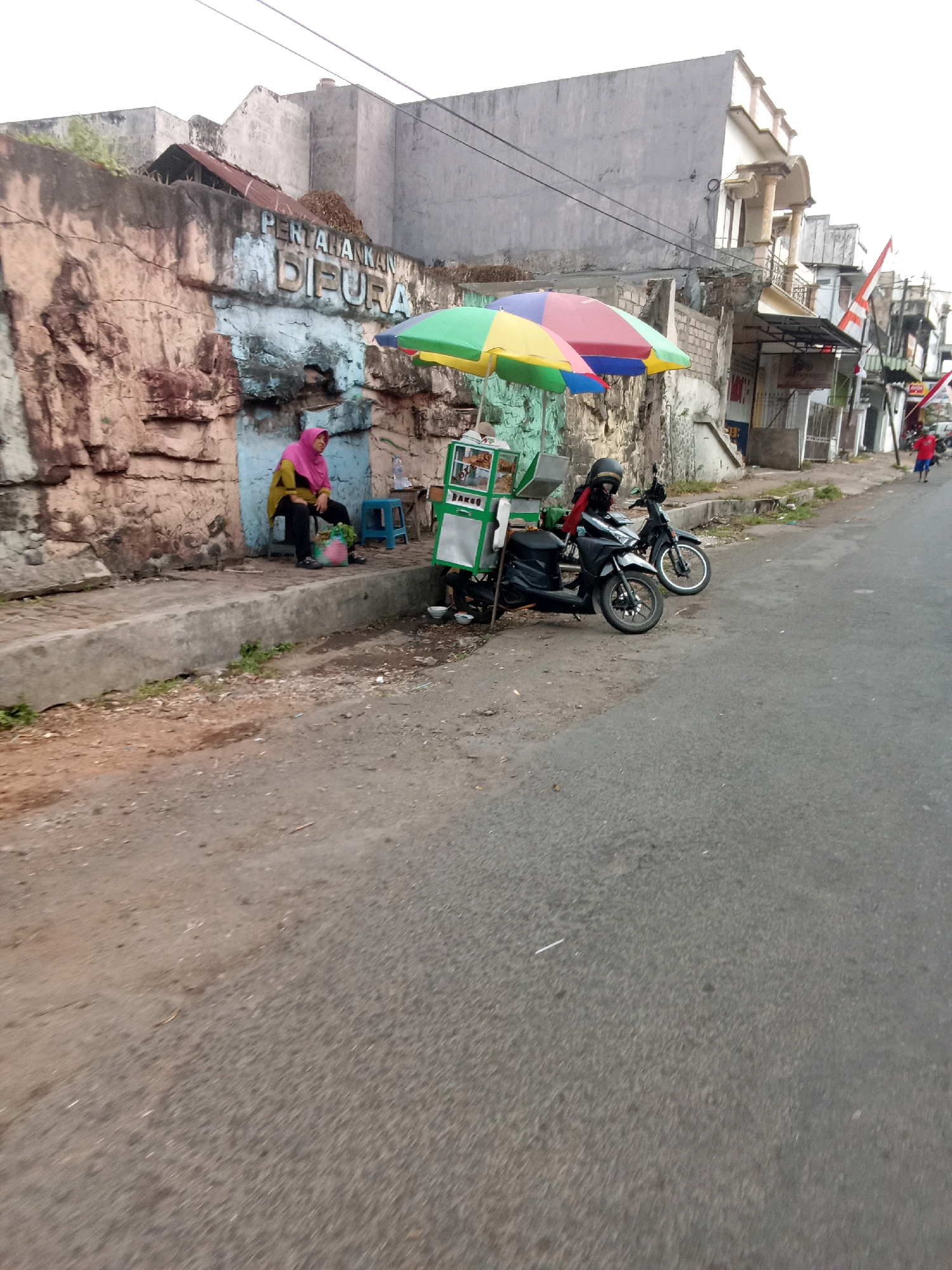 In Jember, Indonesia, a street scene unfolds with a textured, weathered stone wall on the left, featuring the faded inscription "PERTAHANKAN DIFURA." A woman wearing a pink headscarf and yellow top sits on a concrete ledge beside the wall, her legs crossed. Adjacent to her is a bright green food cart, adorned with colorful umbrellas, and two motorcycles parked in front. The street is paved with asphalt, with a dirt shoulder on the left. In the background, buildings with balconies and flags suggest a commercial area. The sky is overcast, and the overall lighting is dim, indicating an early morning or late afternoon.