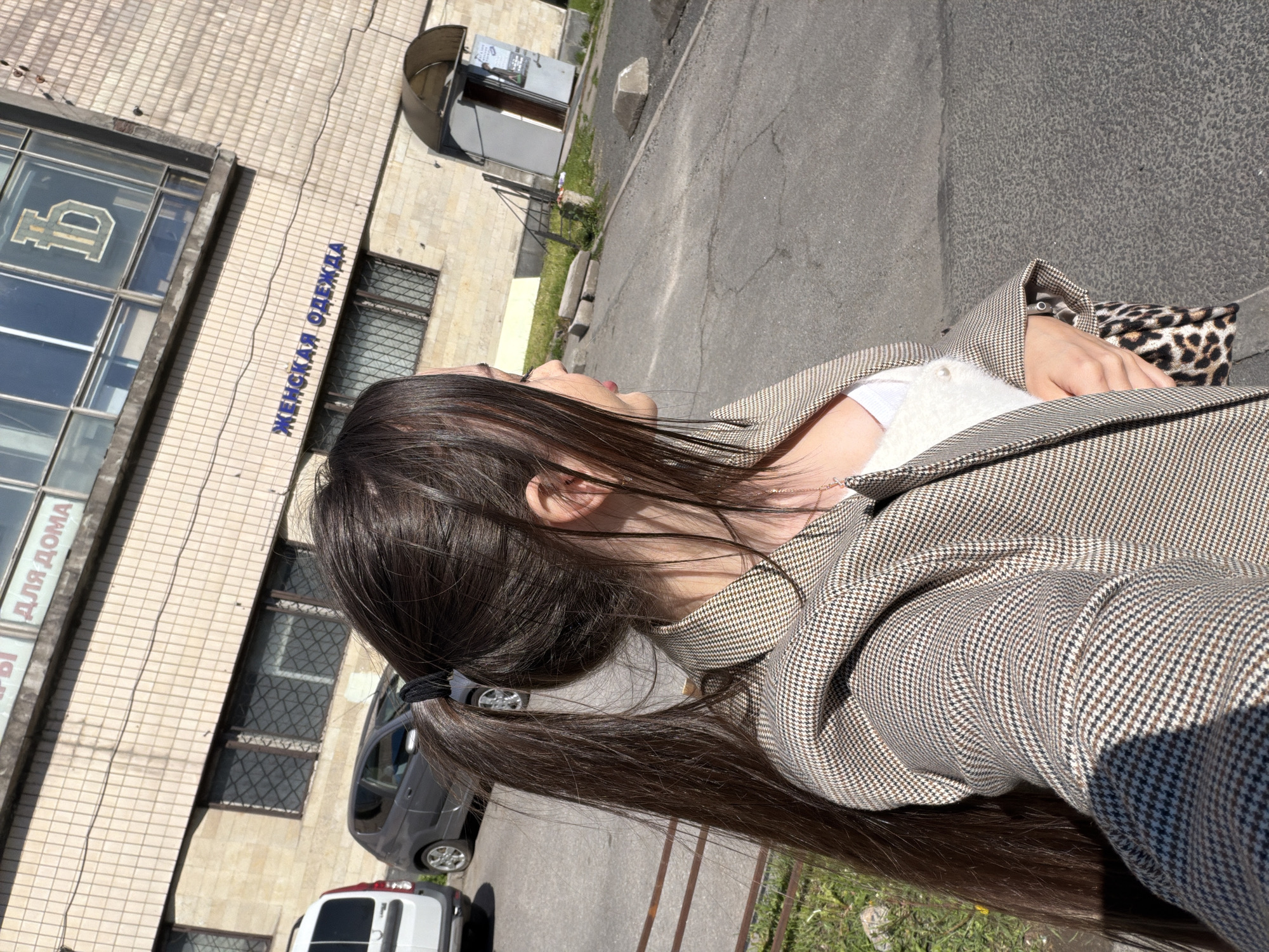 A medium shot shows a woman in Saint Petersburg, Russia. She is wearing a brown-and-white houndstooth jacket over a white top and is holding a leopard-print handbag. She has dark hair pulled back into a ponytail. Behind her, a building with large windows and blue Cyrillic writing is visible. A sign with the ruble symbol is on the window. Parked cars are on the street in front of the building. A gray car and a white van are visible.