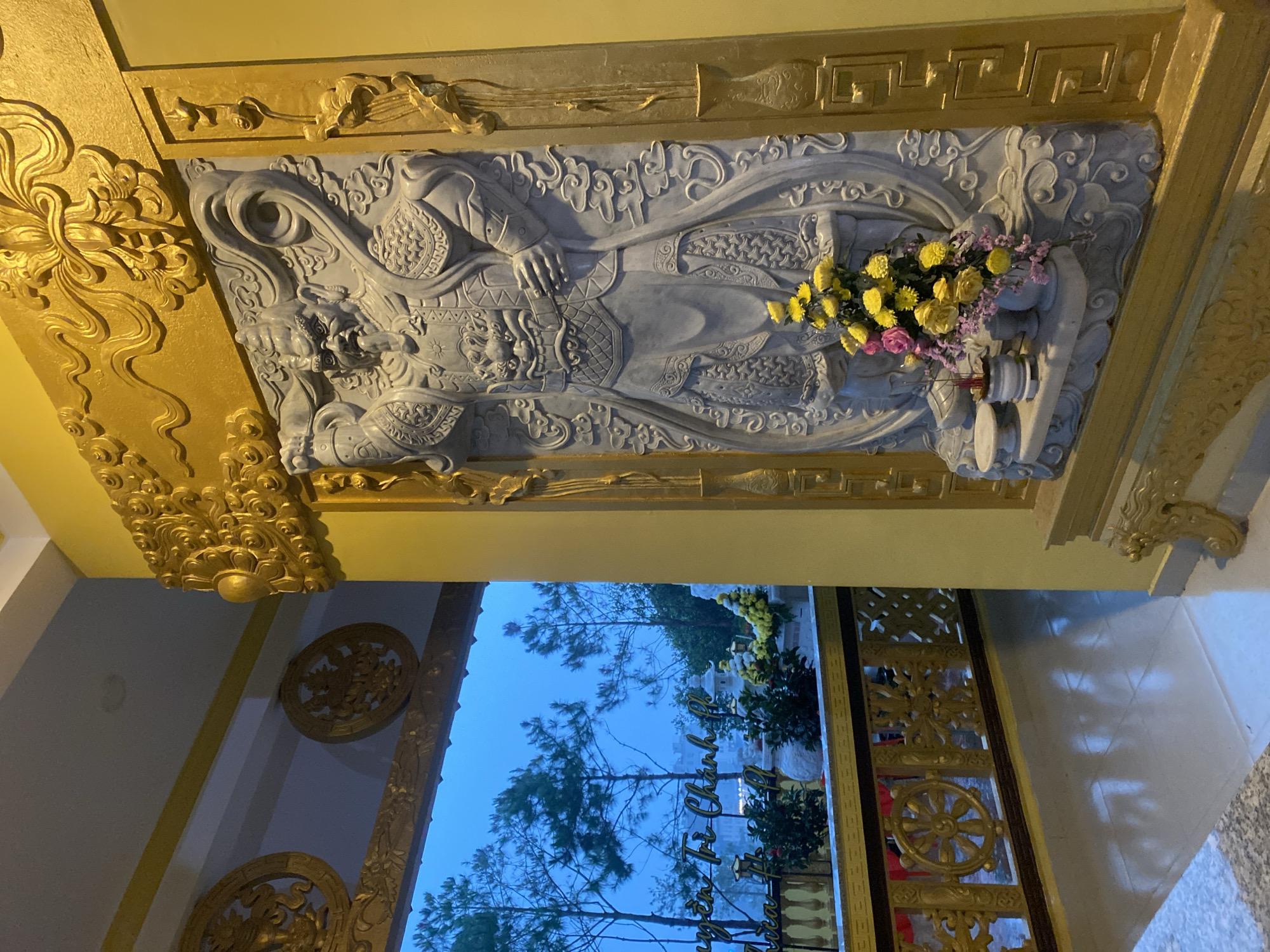 The image depicts the interior of a building, likely a temple or a culturally significant structure, situated in Da Lat, Vietnam.
The focal point is an ornate gray stone carving featuring a recumbent figure, perhaps a deity or a revered person, adorned with flowers. The carving is intricately detailed, with the figure wearing what appears to be armor or traditional clothing. Above it is gold decorative trim.
The building's architecture is visible through a doorway, which provides a view of outdoor scenery with trees and daylight. The building exhibits further elaborate gold ornamentation on the walls and ceiling, indicating a religious or cultural significance. The interior is well-lit, and the colors are rich, adding to the opulence of the space. The overall atmosphere suggests reverence and cultural heritage.