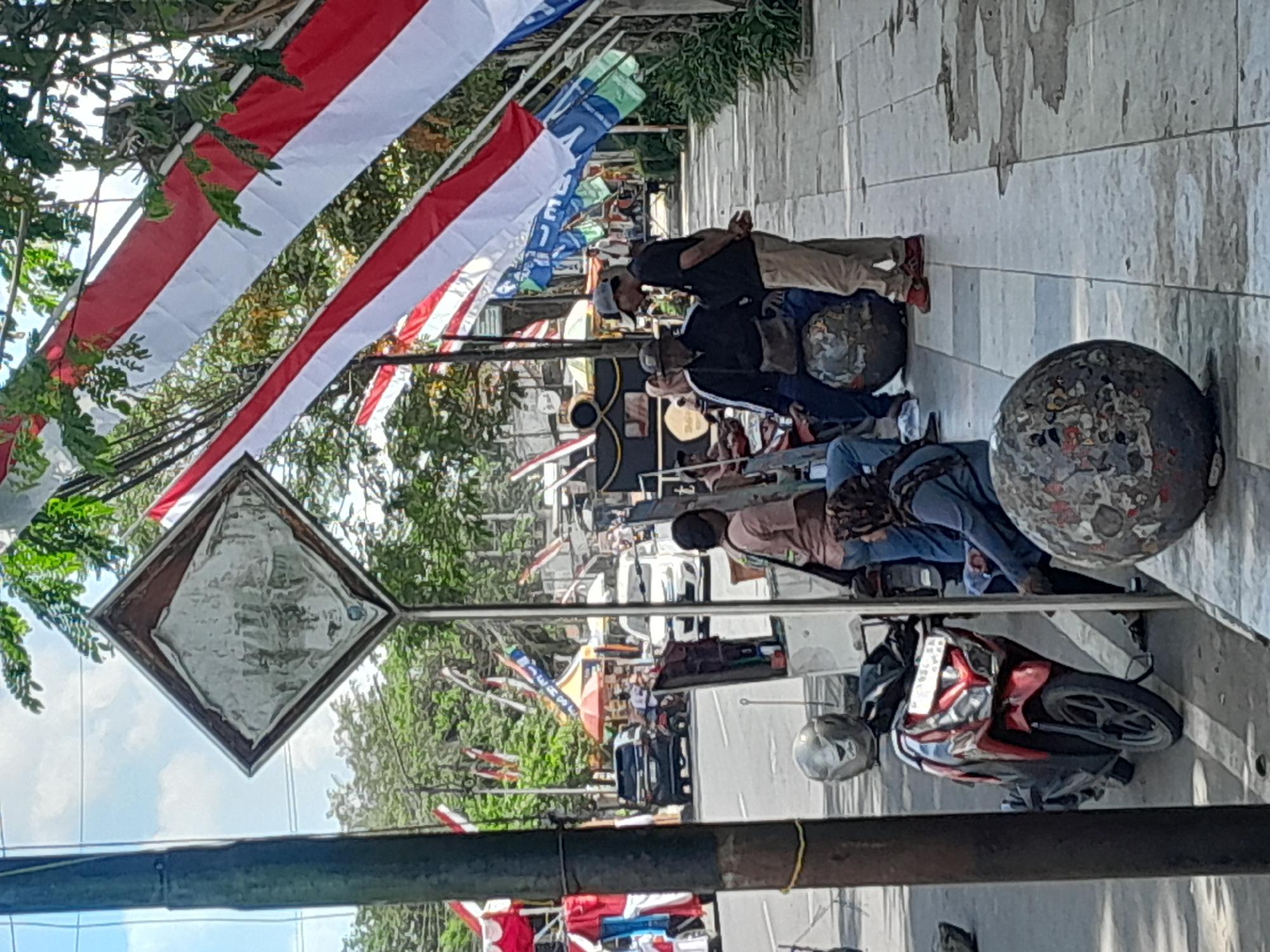 The image captures a street scene in Genteng Kulon, Indonesia, characterized by vibrant Indonesian flags and a lively pedestrian area. In the foreground, a red and black scooter is parked. To the right, three individuals are gathered on a sidewalk. One person, wearing a cap and dark shirt, stands and converses with another individual seated on a large, spherical object. A third person, also seated on a similar spherical object, is visible to the left of the standing figure. Further into the background, the street is busy with vehicular traffic, including a black SUV and a white car. Numerous stalls and awnings are set up along the roadside, decorated with Indonesian flags and banners, suggesting a festive or market atmosphere. Trees and foliage line the street, providing a green backdrop to the urban environment. A diamond-shaped signpost stands prominently on the left side of the image. The overall scene conveys a sense of daily life and community engagement in the Indonesian city.
