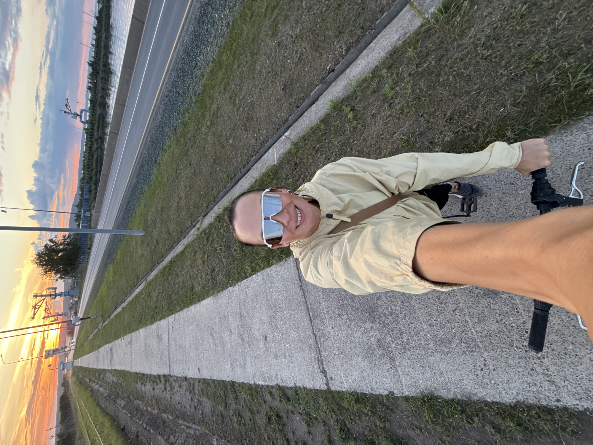 The image shows a person on a bicycle, taking a selfie. The person is wearing a light-colored jacket and white sunglasses. They are smiling and appear to be enjoying themselves. The bicycle's handlebars are visible in the foreground, and the person's arm is extended to hold the camera.
The setting is an outdoor path next to a road. There is a strip of grass between the path and the road. In the background, a body of water is visible, possibly a river or a bay, with industrial structures and cranes on the far side. The sky is filled with colorful clouds, suggesting either sunrise or sunset. The overall atmosphere is serene and peaceful.
The time of day appears to be either early morning or late evening due to the warm hues in the sky. The weather seems pleasant. The presence of a road and industrial elements might suggest the location is near a port or an industrial zone. The image is taken in Chaikovsky, Russia. No visible text is present in the image.
