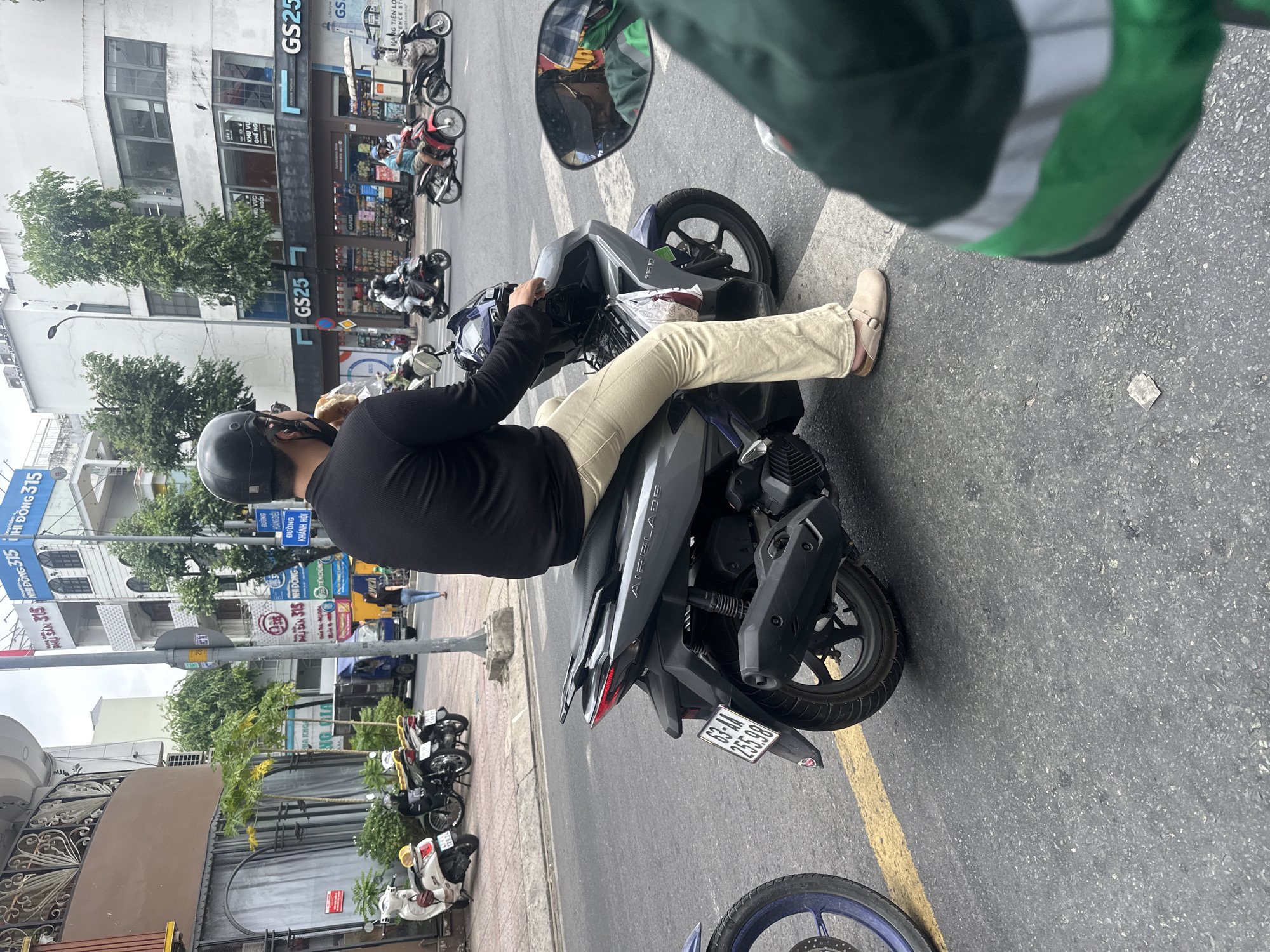 This image depicts an urban street scene in Thủ Đức, Vietnam, under an overcast sky. In the immediate foreground, to the right, is the blurred shoulder and upper arm of an individual wearing a green and white uniform, likely the photographer or a passenger on an adjacent motorbike. The reflection of this individual, wearing a cap and dark glasses, is visible in the right rearview mirror of the primary subject's motorcycle. A blue motorcycle wheel is partially visible on the far left foreground.
The central subject is a male individual, observed from behind, seated on a grey Honda Airblade 150 scooter with license plate 63-AA 255.98. He is wearing a black full-sleeved shirt, beige trousers, and light-colored slip-on shoes. A black full-face helmet and dark sunglasses are worn. His right hand holds a baguette-like bread item, positioned near his mouth, indicating consumption. The scooter is stopped on asphalt pavement marked with a yellow lane line.
In the midground, multiple motorbikes are visible on the road, with several riders wearing helmets, suggesting active traffic. Beyond the traffic, the background consists of multi-story commercial buildings. To the right, a large white building houses two "GS25" convenience stores, with additional signage for a "children's play area" and "convenience goods." Trees are planted along this building. To the left, a more ornate building with a curved, brown facade and large windows is visible, with smaller palm trees in front. Further left, another building displays Vietnamese signage, including "PHÚ SÂN 315" and "NHỊ ĐỒNG 315." A blue street sign reading "ĐƯỜNG KHÁNH HỘI" is partially visible. A blue commercial truck is parked on the street in the distance on the left. Sidewalks on both sides of the street contain parked motorbikes, and one individual is seen walking on the left sidewalk. Utility poles and overhead wires are present throughout the background.