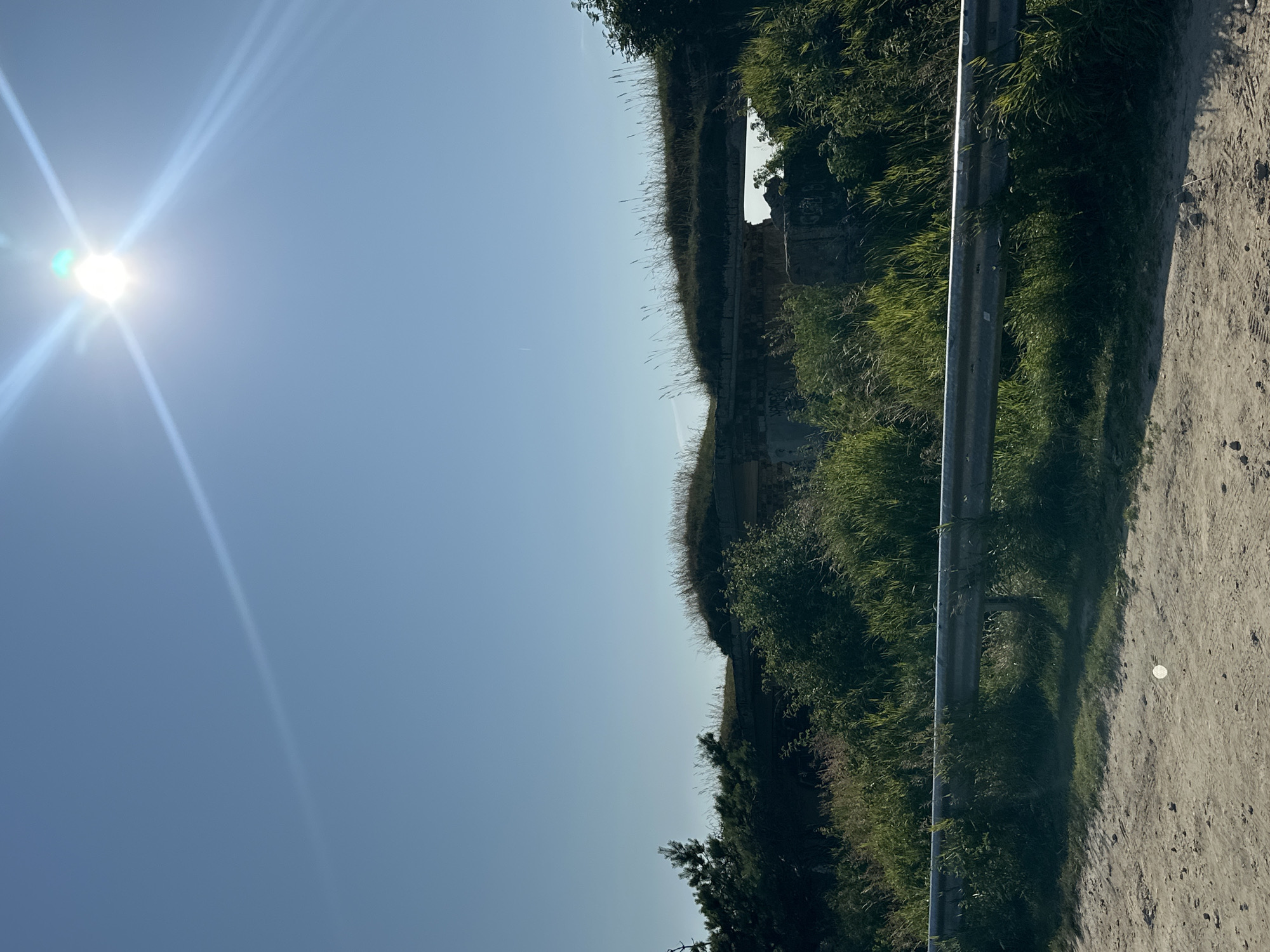The image depicts a sunny day in Liepaja, Latvia, with a clear blue sky. The foreground shows a dirt road with a metal guardrail running horizontally across it. Beyond the guardrail, lush green grass and vegetation grow, partially obscuring an old, overgrown concrete structure. This structure appears to be part of a historical fortification or bunker, with grass growing on its roof and openings visible. The sun is high in the sky, creating lens flare and bright light beams. The overall scene suggests an outdoor, possibly rural or coastal, environment.