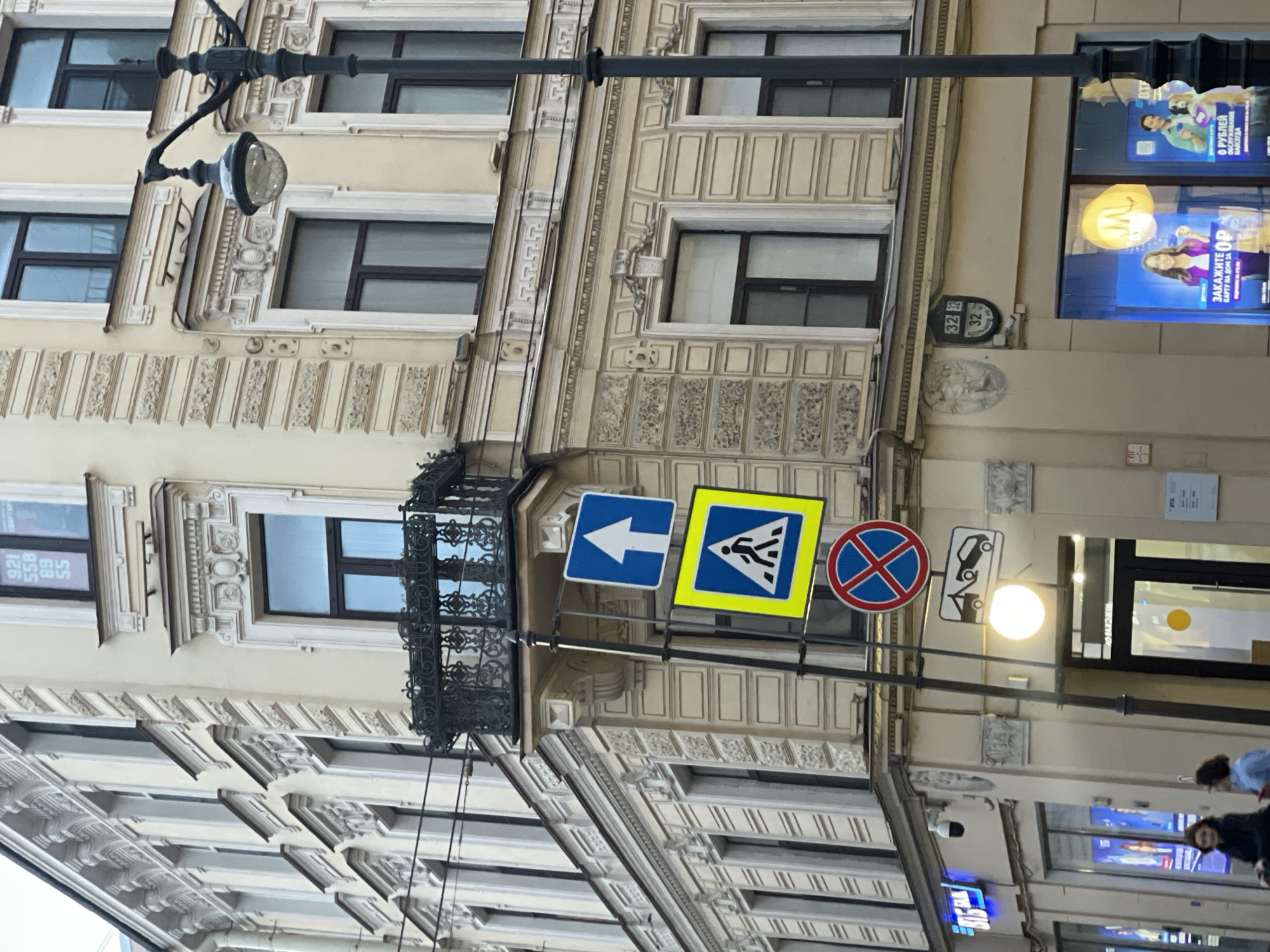 The image depicts a street scene in Saint Petersburg, Russia. The focus is on the architectural details of a building facade, showcasing elements like windows, decorative moldings, and a wrought-iron balcony. Several street signs are attached to a pole near the building, including a blue arrow indicating a direction, a pedestrian crossing sign, and a "no stopping" sign. Additionally, a street lamp is visible. The background features advertising posters displayed in the windows of a business. Several people are walking on the sidewalk. The OCR identifies numbers 921, 558, 89, 55, 32, 32, 32, and some Russian text.