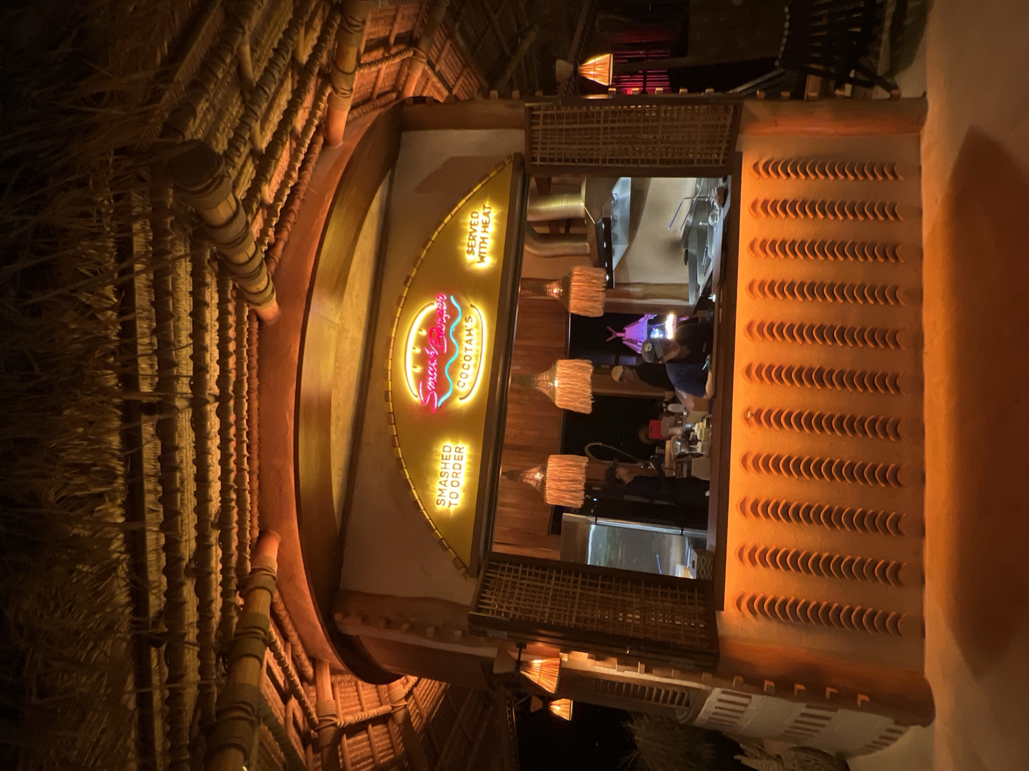 A brightly lit, open-air food stall is depicted under a thatched roof structure at night. The stall, likely located in Baan Bo Phut, Thailand, features a neon sign above the counter that reads "SMASHED TO ORDER" and "Smash Burger COCOTAM'S SERVED WITH HEAT". The counter area is illuminated by warm orange lights, highlighting a textured wall with a series of semi-circular cutouts. Several employees are visible behind the counter, some appearing to be preparing food. Three decorative lamps with fringe hang from the ceiling. Wooden shutters frame the service window. The overall atmosphere suggests a casual dining establishment.