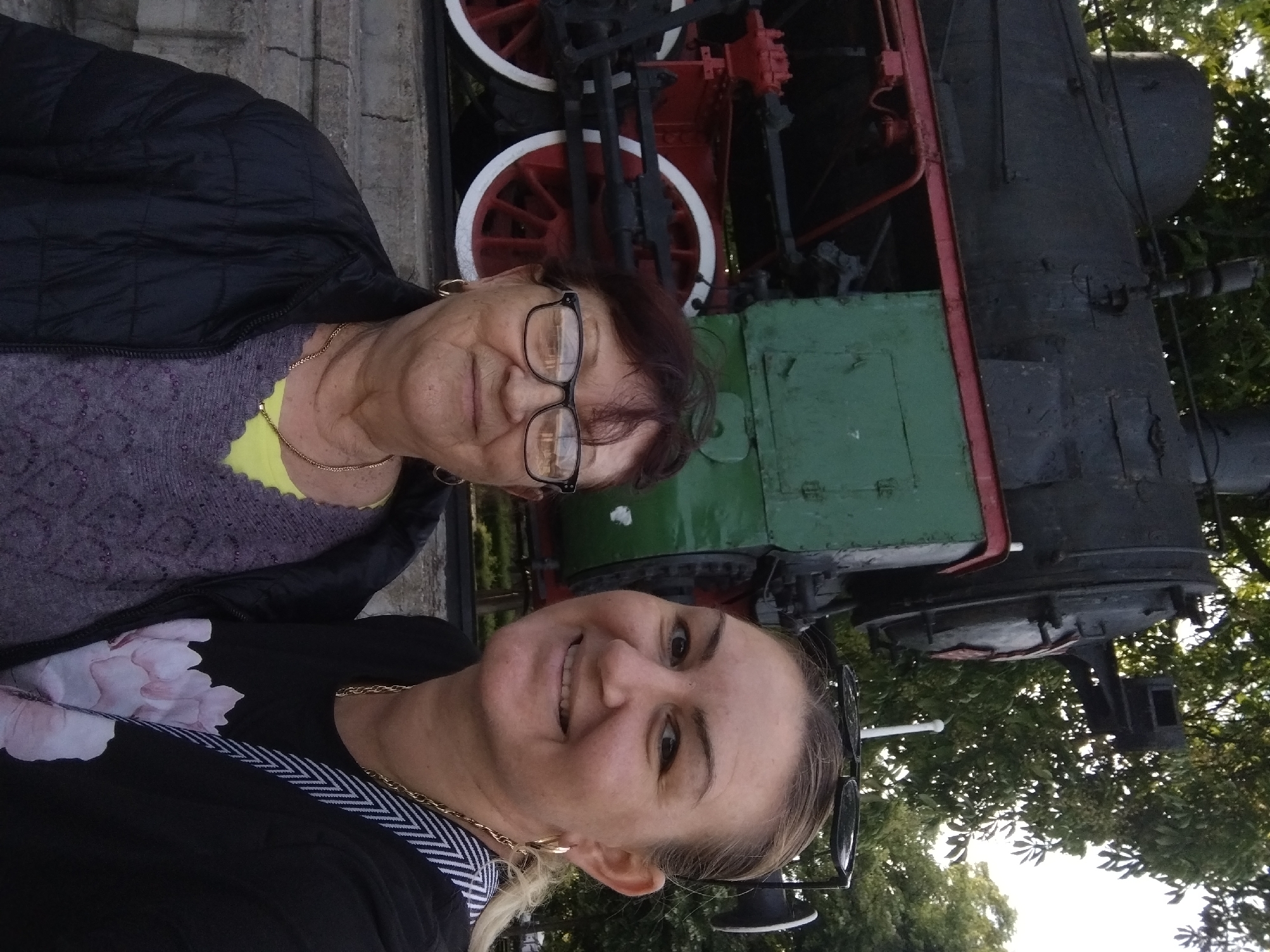 Two women pose for a photo in front of a vintage steam train at a location most likely in the city of Stara Zagora, Bulgaria. The older woman wears glasses, a black jacket, and a patterned sweater. The younger woman is smiling, wearing a black top with a floral design and a gold necklace. The steam train behind them is painted in red, green, and black. The background includes trees, suggesting an outdoor or park setting.