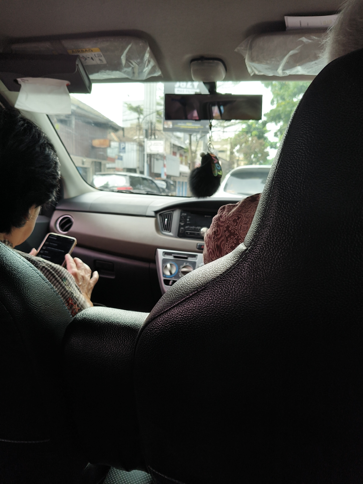 The image is taken from the back seat of a car, looking forward. Two people are visible. In the left foreground, a person with dark hair is holding a smartphone and appears to be using it. They are wearing a checkered shirt over another garment. To the right of this person and further into the background, another person is visible, wearing a patterned garment. They are looking forward. The interior of the car includes a dashboard with air vents and a radio or media console. A rear-view mirror is visible overhead, with a fuzzy black charm hanging from it. Through the front windshield, a street scene is visible, with buildings and trees. The overall impression is that the car is in motion within an urban environment. The specific location in the city of Bandung, Indonesia cannot be determined from the visual information provided.