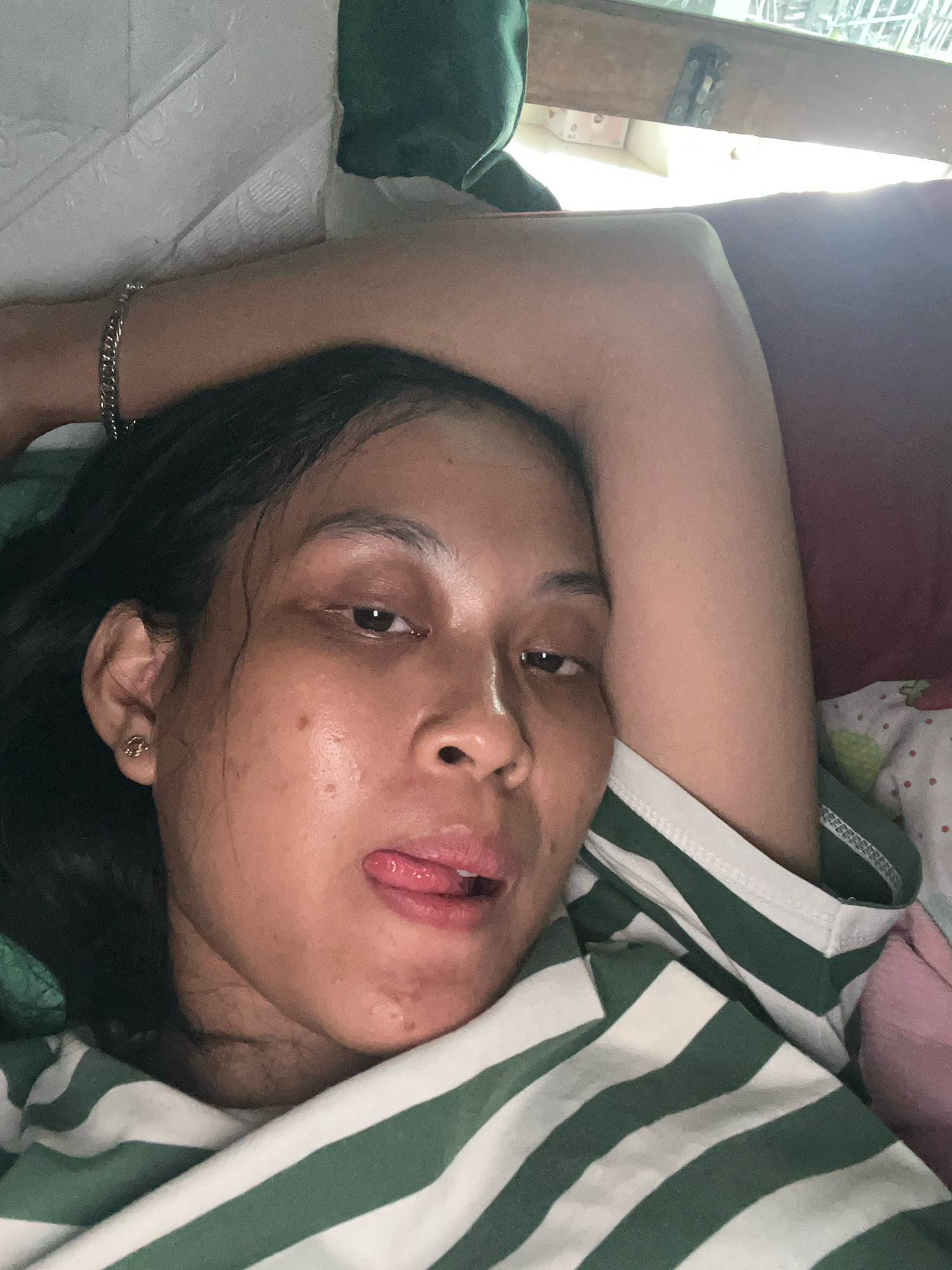 A close-up, eye-level shot features a young woman lying down, her head resting on her arm. Her eyes are open, and her tongue is sticking out slightly. She is wearing a green and white striped shirt. The background includes what appears to be bedding and a window with sunlight filtering through. There are no discernible landmarks or features that would indicate a specific location within Kecamatan Pati, Indonesia.