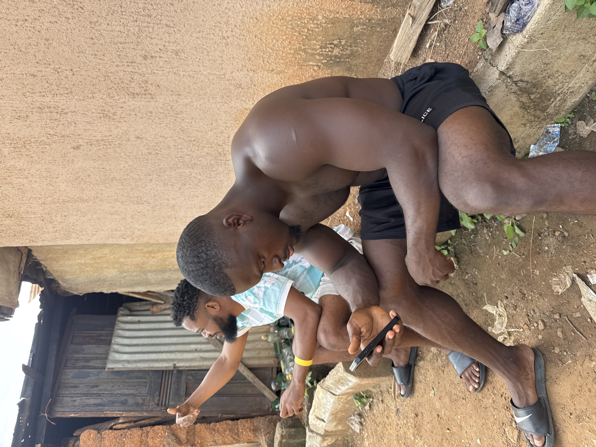 Two Black men are seated outdoors in a gritty urban environment, possibly in Akwanga, Nigeria. The man in the foreground, shirtless and wearing black shorts, is looking down at his smartphone which he holds in his right hand. His left arm rests on his lap. He has muscular build. The man behind him, wearing a light-colored t-shirt with a blue and white pattern and white shorts, is looking to his left with his left arm raised in a thumbs-up gesture. He has a beard and is wearing a yellow wristband on his left wrist. They are seated on the ground next to a concrete barrier. Behind them are elements of informal construction: a corrugated metal sheet, a wooden door, and walls made of reddish-brown material and a rough, beige stucco. Scattered on the ground are plastic bottles and debris, typical of an outdoor, less-maintained area. The lighting suggests it is daytime.