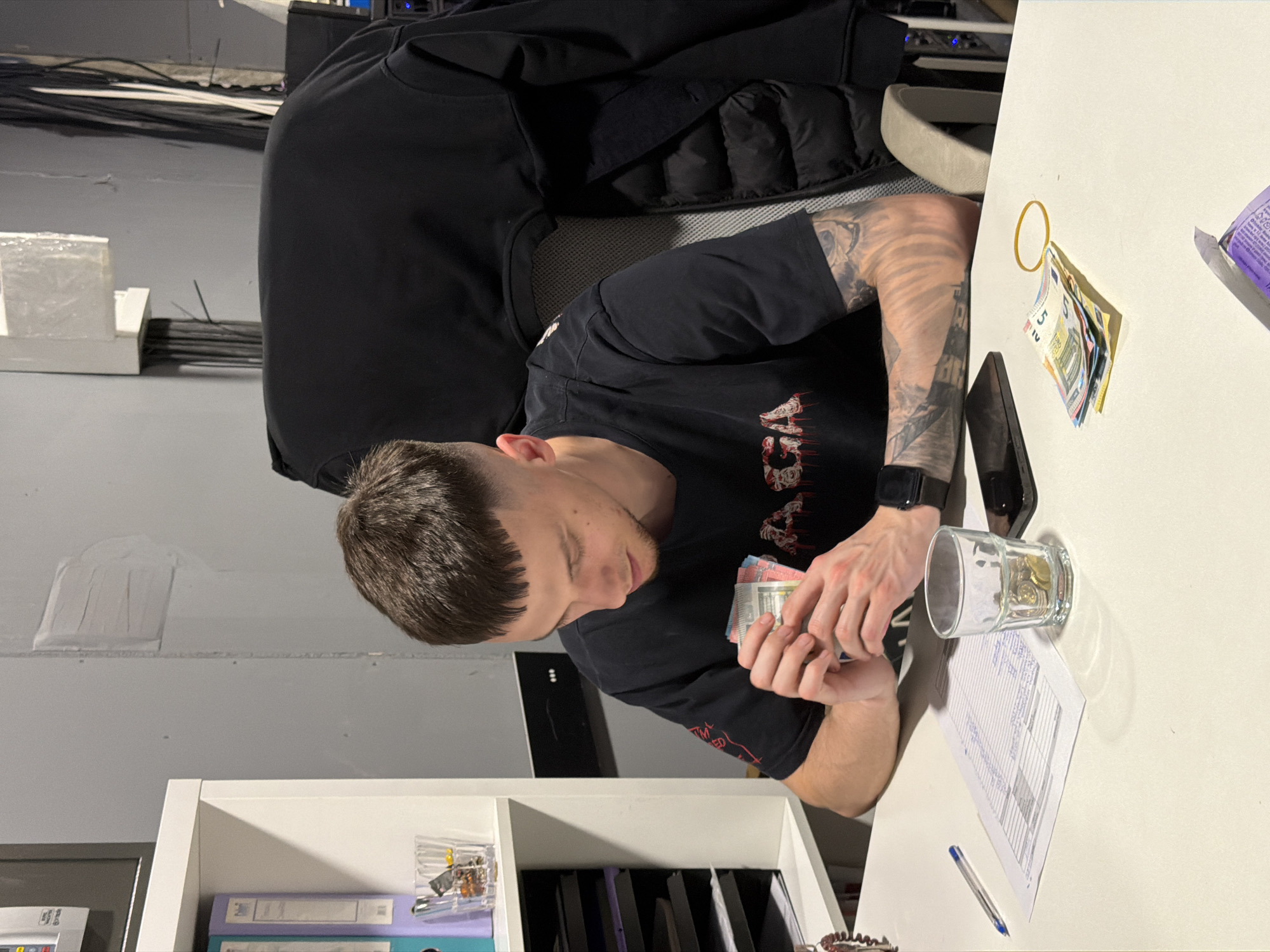 A man is seated at a white desk in an indoor environment. He is counting a stack of bills. A glass jar containing coins rests on the desk next to a printed document and a pen. A cell phone and another bundle of bills secured with a rubber band are also visible on the desk. A black jacket is draped over the back of the chair. A white shelving unit is underneath the desk. The location cannot be determined as Varna, Bulgaria from the image.