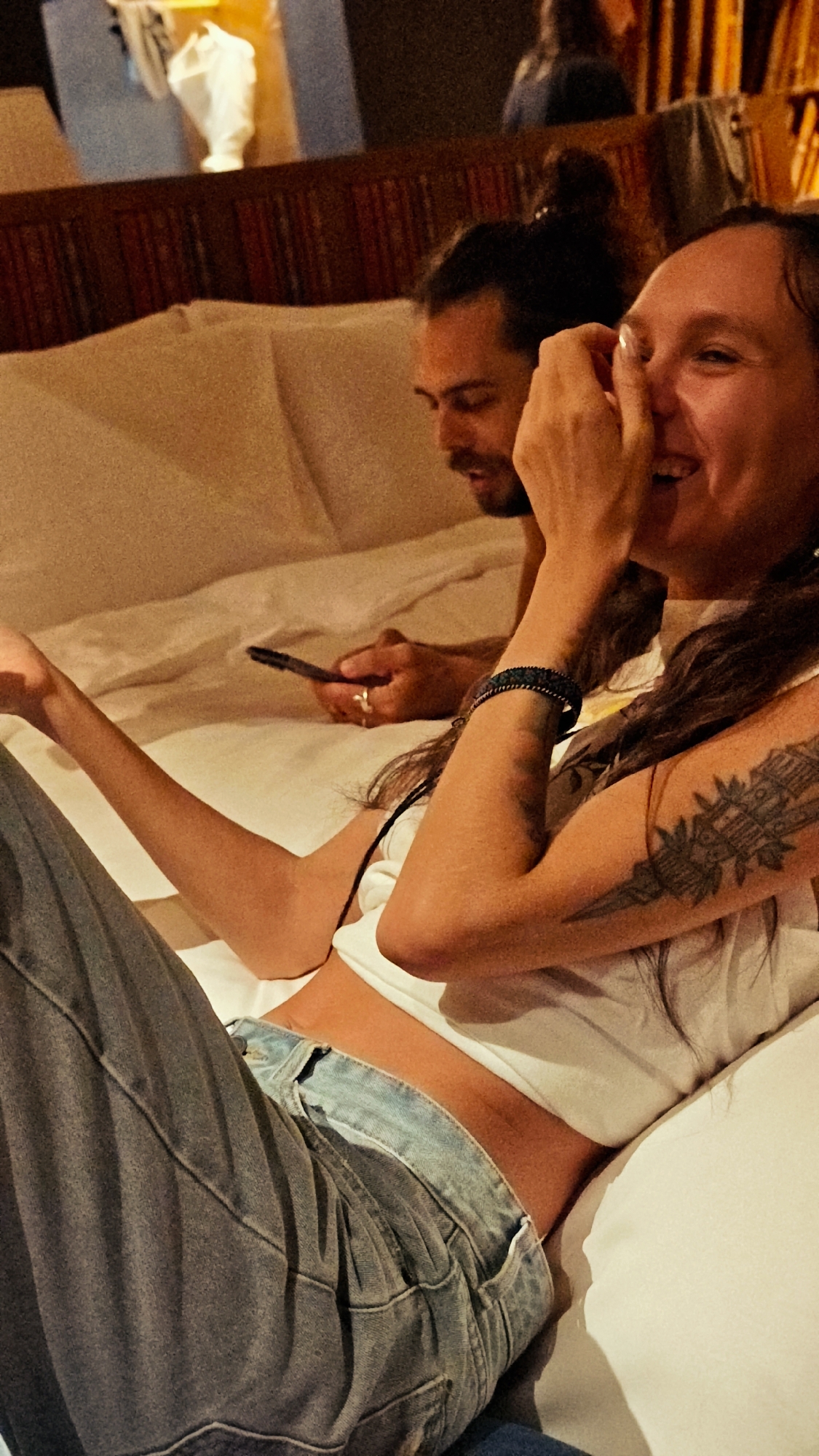 In Mù Cang Chải, Vietnam, an indoor scene depicts two individuals relaxing on a bed. In the foreground on the right, a woman with long dark hair, wearing a white sleeveless crop top and blue jeans, smiles broadly with her right hand covering her mouth. A dark, geometric tattoo is visible on her left upper arm, and a dark woven bracelet is on her left wrist. Her left arm rests on the white bedding. Behind her to the left, a man with dark hair tied in a bun and a beard looks down at a black smartphone held in his hands. He wears a light-colored top, and a ring is visible on his right ring finger. The bed is made with white sheets and pillows. In the background, a dark wooden headboard or wall paneling is present, and further back, blurred elements suggest additional furniture or items and soft, warm lighting.