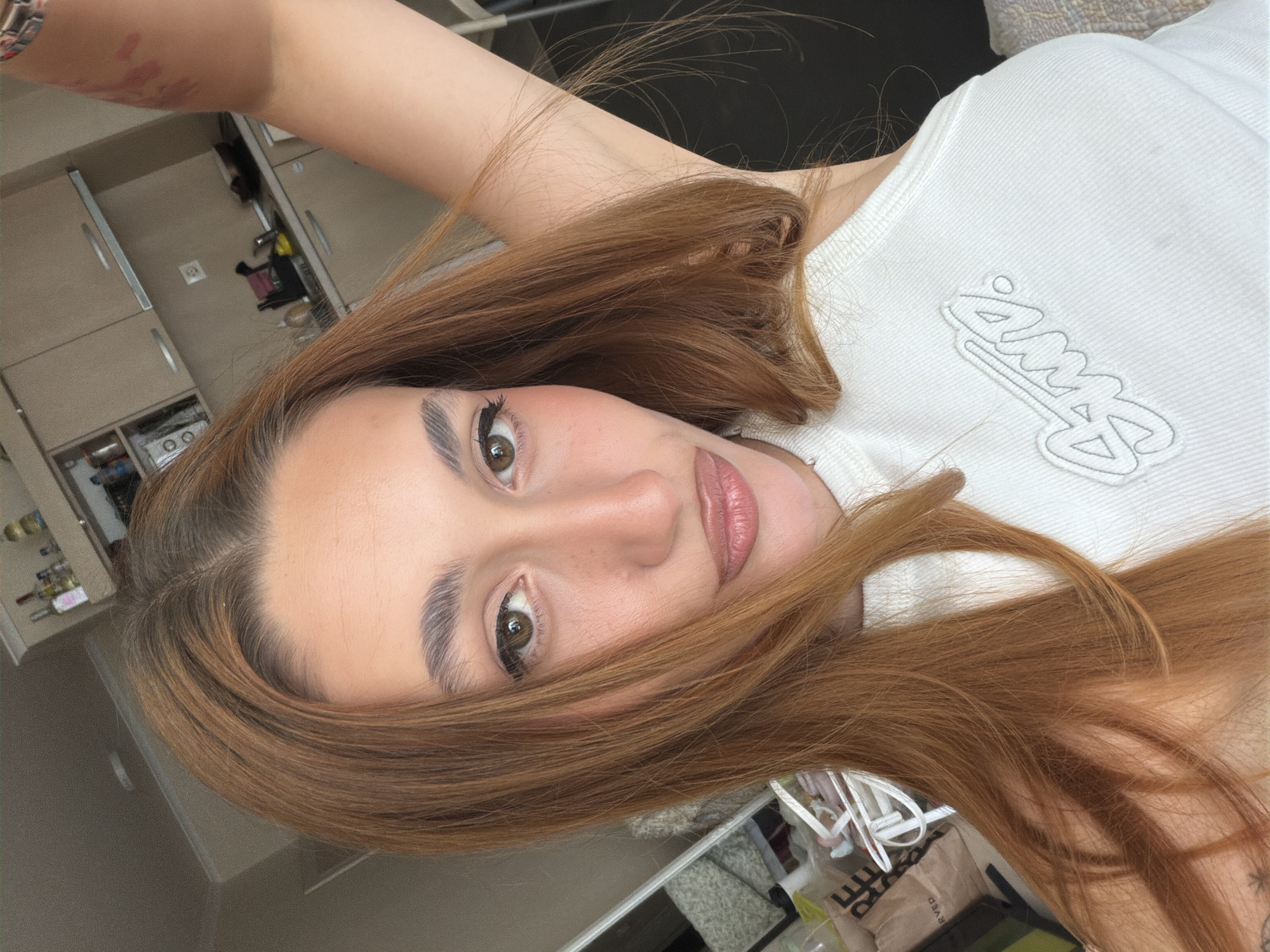 A young female, with long reddish-brown hair parted off-center, is depicted in a close-up selfie-style photograph. She has prominent eyebrows, wears winged black eyeliner, and light lip gloss, with visible freckles on her nose. Her expression features slightly pursed lips. She is wearing a white ribbed tank top with a light grey embroidered "SMW" logo on the chest. Her right arm is raised, presumably holding the camera, revealing a bare shoulder and upper arm with several faint, linear pink marks on the bicep/tricep area.
The background shows an indoor domestic setting, likely a kitchenette or apartment. To the right, there are light brown cabinets with silver bar handles, a kitchen sink with a faucet, and various bottles and objects on the counter, including a yellow item resembling a rubber duck. An electrical outlet or switch is visible on the wall. To the left, a partial reflection in a mirror reveals elements of the room, including a brown bag labeled "RESERVED."
The scene is situated in the city of Sofia, Bulgaria.