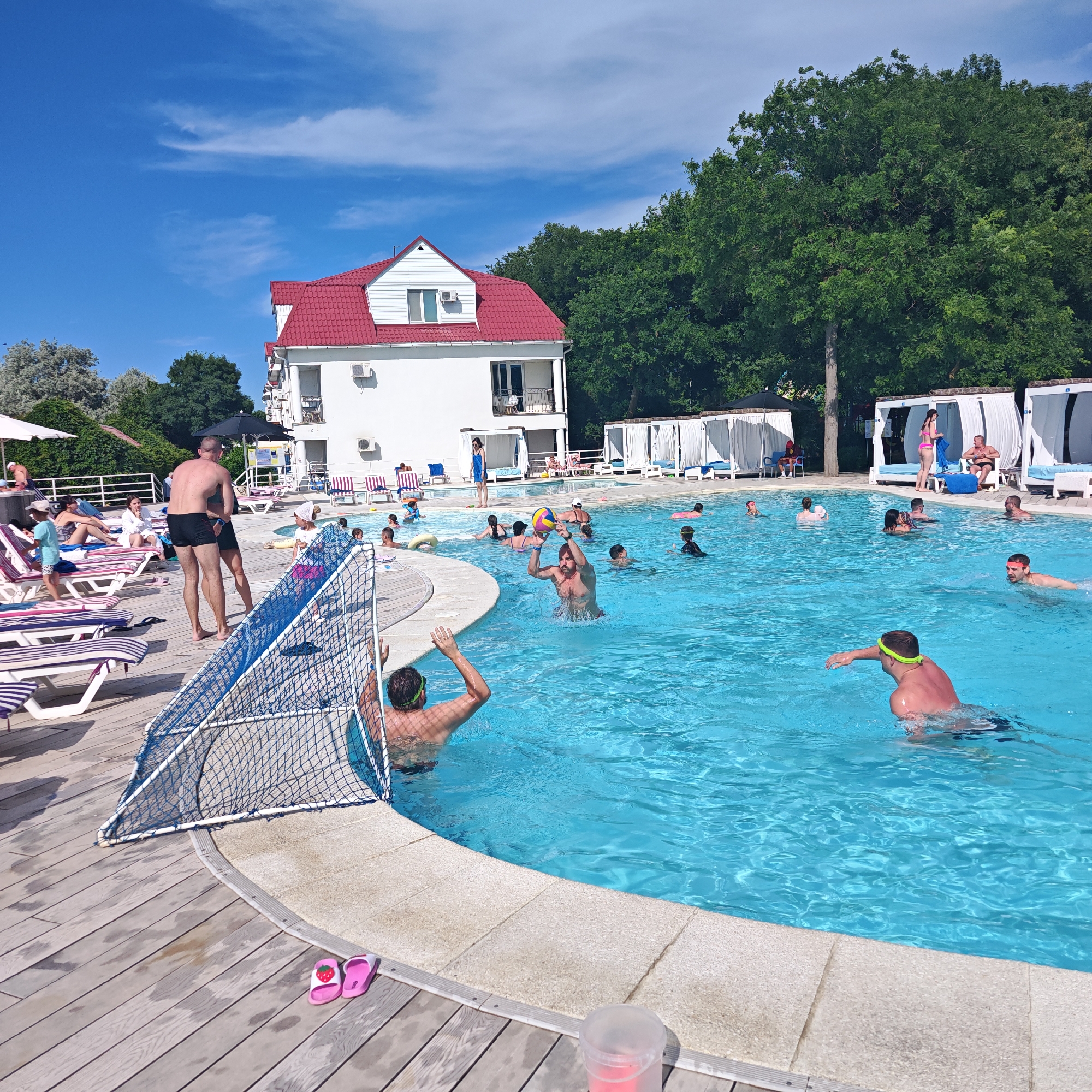 In a bright, sunlit outdoor swimming pool, several people are engaged in water activities. The setting appears to be a resort or hotel, with lounge chairs and cabanas lining the wooden deck surrounding the pool. A white building with a red roof stands in the background, partially obscured by lush green trees. The sky is a clear blue with a few wispy clouds.
In the pool, a group of individuals are playing a water game, possibly water polo, with a colorful ball. One man is holding the ball aloft, seemingly in the midst of a throw or catch. Another man in goggles is swimming towards the net on the left side of the image, which is set up for the game. Several other people are scattered throughout the pool, some swimming, others socializing, and one person appears to be wearing a flotation device.
On the deck, a few people are lounging on chairs, while others are walking or standing. Two pairs of flip-flops, one pink with a strawberry design and another plain pink, are placed on the wooden deck near the pool's edge. The overall atmosphere is relaxed and joyful, suggesting a warm, sunny day during a vacation or leisure time. The scene is set during daylight hours, likely midday given the bright sunlight. There is no visible text in the image.