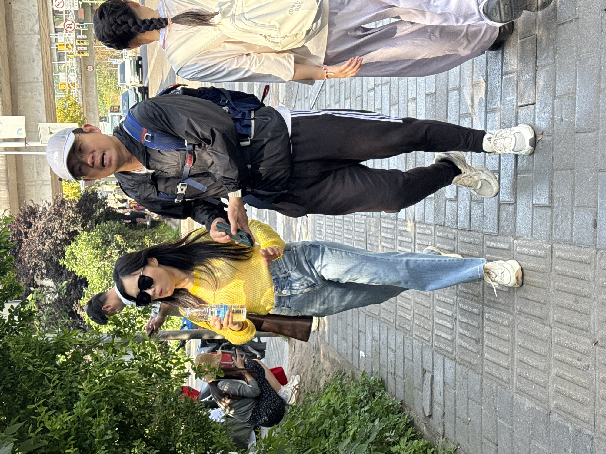 The image depicts an urban sidewalk scene in Nan'an District, China, on a bright, sunny day. Several individuals are visible, with three prominent figures in the foreground and midground.
In the midground, a man wearing a white baseball cap, a black jacket, and black track pants with white stripes (reminiscent of Adidas) stands on the paved sidewalk. He has a blue backpack and his mouth is open as if speaking or reacting. To his left, a woman with long dark hair wears sunglasses, a bright yellow long-sleeved top, blue jeans, and white sneakers. She holds a clear plastic water bottle in one hand and a phone in the other, looking downwards or to her side.
Behind the man, a third person is partially visible, dressed in light-colored clothing, including a white top and grey pants. They have dark hair styled in a braid and carry a light-colored backpack with the text "KDMILL BAG" partially legible. Further in the background, other individuals are partially obscured by lush green bushes and trees lining the sidewalk. One person is seen sitting near the bushes, wearing a straw hat.
The background reveals a multi-lane road with cars, and several traffic light poles. A large concrete structure, possibly an overpass or elevated road, spans across the upper part of the background. The sidewalk is constructed from grey paving stones laid in a geometric pattern. The overall scene suggests a casual day out, with people walking and standing in an active urban environment.