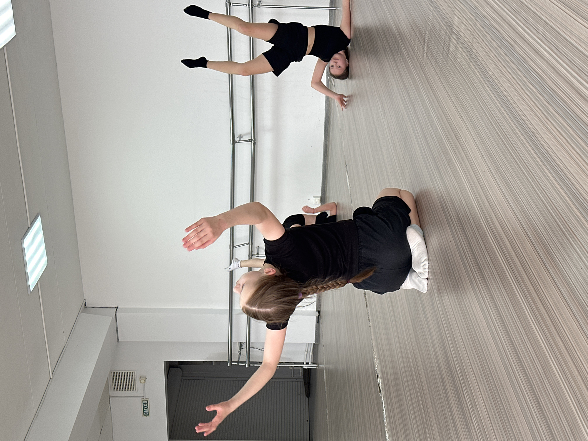 Two young female dancers are in a dance studio. The dancer in the foreground is kneeling with one knee on the floor and her arms extended. She has long hair in a braid. She is wearing a black shirt and black shorts. The dancer in the background is doing a handstand with her legs extended. She is wearing a black top and black shorts, and black socks. The dance studio has a white wall with a ballet barre and a light gray wooden floor. The studio is located in the city of Votkinsk, Russia.