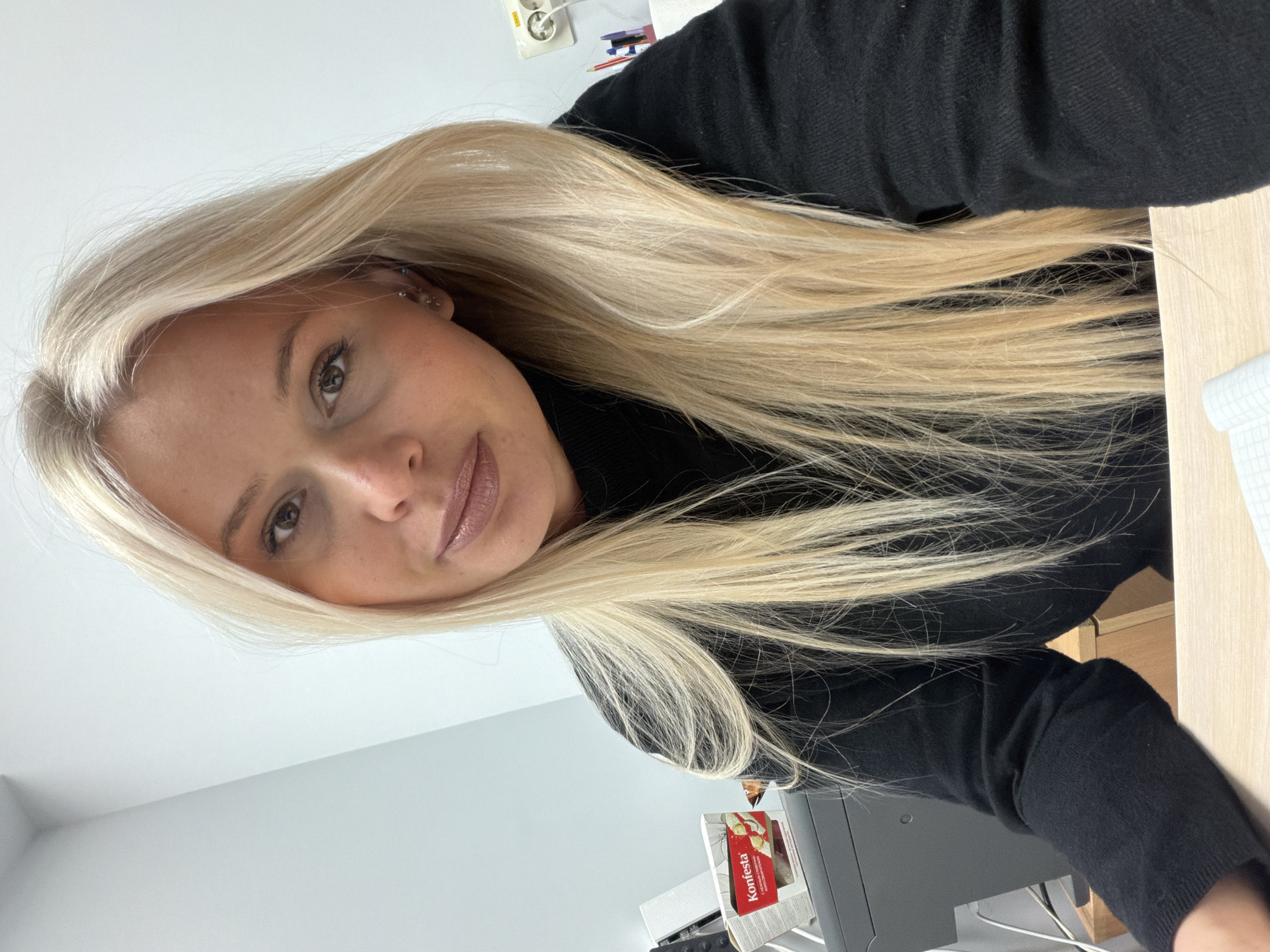 A woman with long, light blonde hair is posing for a selfie. She has fair skin, brown eyes, and is wearing a dark-colored turtleneck sweater. She appears to be indoors in an office setting. A desk is visible to her right, and a printer and box of "Konfesta" sweets are in the background. The image location is inferred to be in Sarapul, Russia, based on the branding of the box.