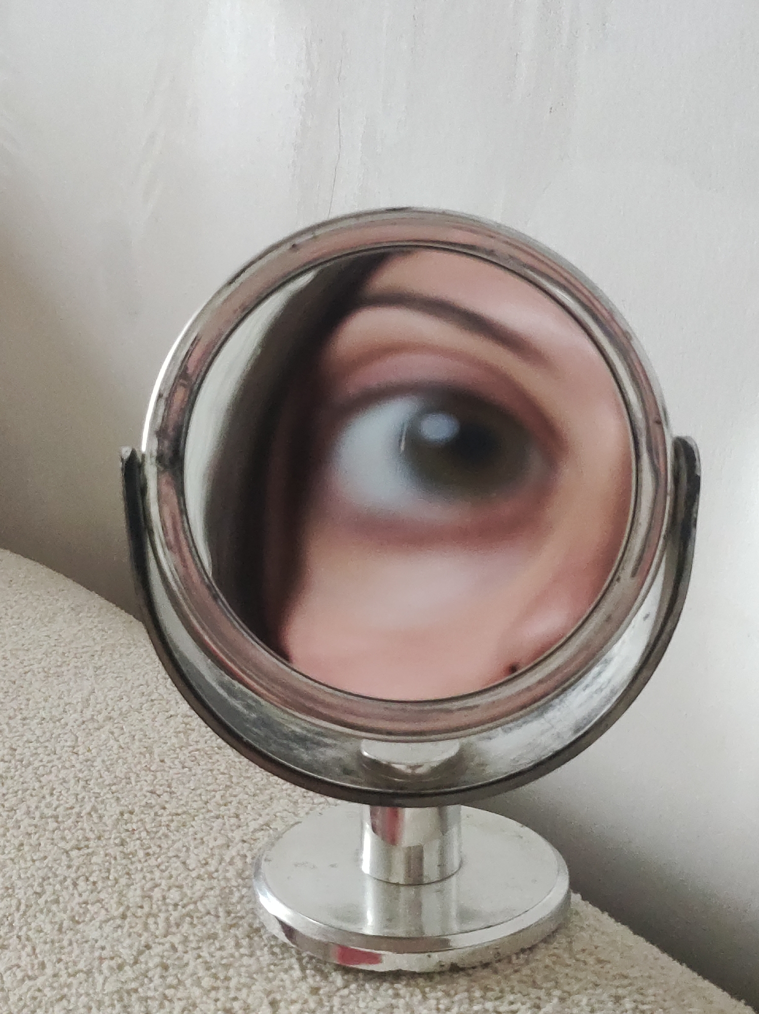 A close-up, indoor shot depicts a small, round, metallic makeup mirror reflecting a human eye. The eye appears to be brown or hazel. A portion of the cheek and brow area is also visible in the mirror's reflection. The mirror has a circular base and support arms, and rests on a light-colored, textured surface. The background appears to be a plain white or off-white wall. The location is not discernible from the image.