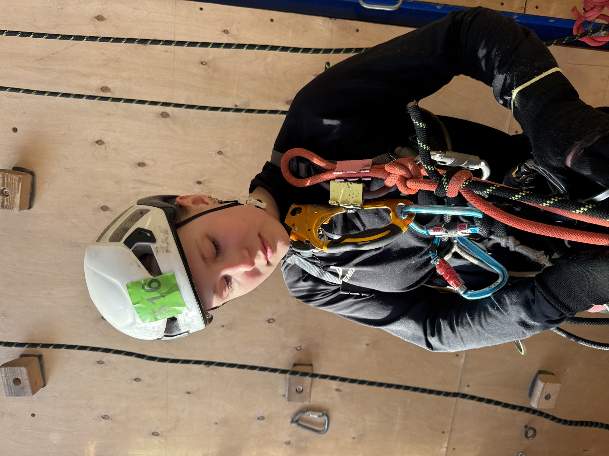 A person is depicted in an indoor climbing environment. The individual is wearing a white helmet with a green sticker, a black long-sleeved shirt, climbing harness, and gloves. They are equipped with various climbing equipment, including ropes, carabiners, and a belay device. The background consists of a wooden climbing wall with installed climbing holds and ropes. The image does not contain enough data to determine the location of the depicted person.