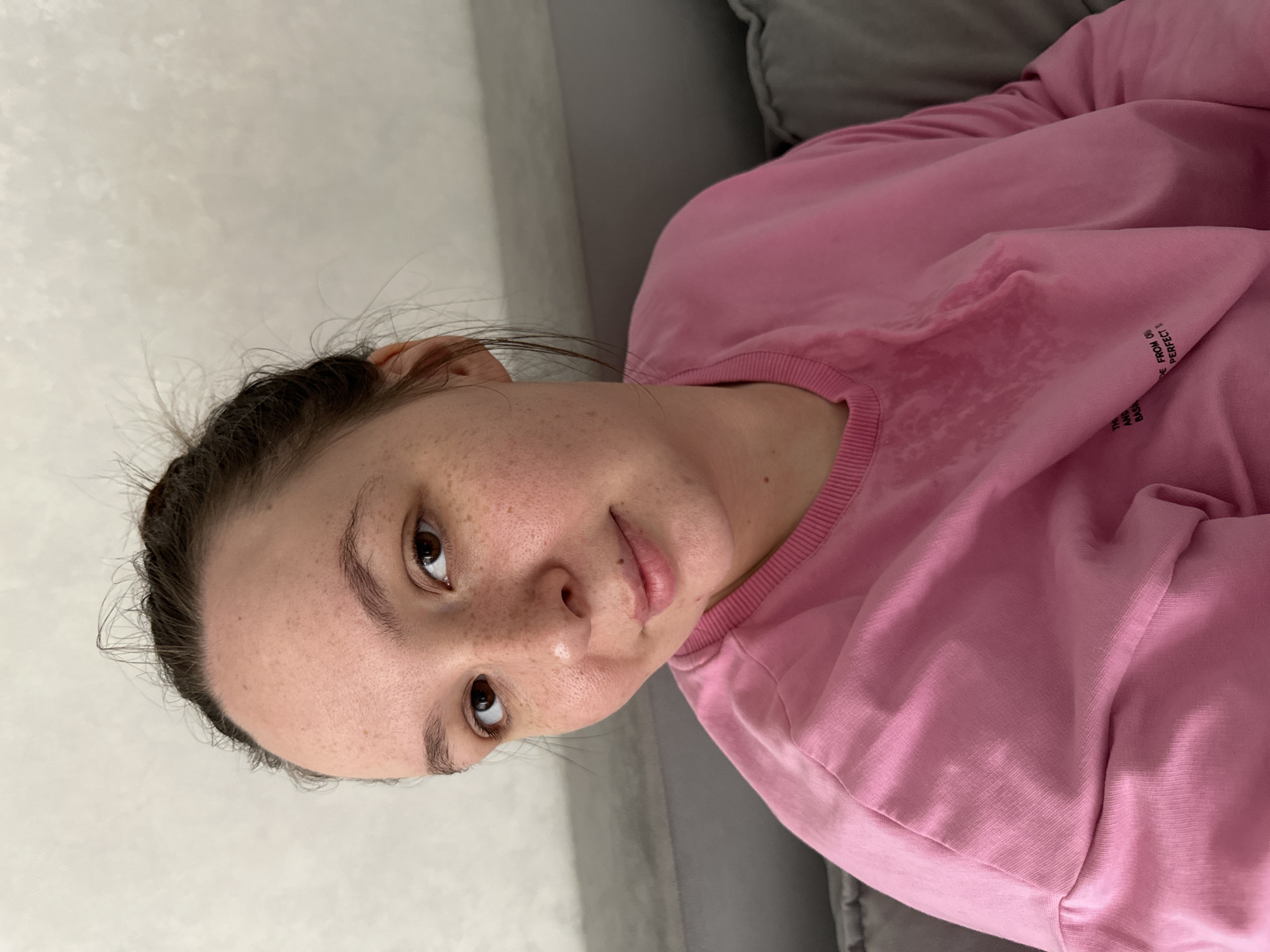 A young woman with brown hair pulled up into a bun and freckles across her face is shown in a close-up, head-and-shoulders shot. She is wearing a pink sweatshirt with a slightly damp area on the chest. Her hair is slightly messy, with strands falling around her face. She is looking directly at the camera with a slight smile. The background appears to be an indoor setting with a textured light-colored wall and a portion of a gray couch visible on the left and right sides of the frame. There are no identifiable landmarks or clues to suggest the specific location within Chaikovsky, Russia.