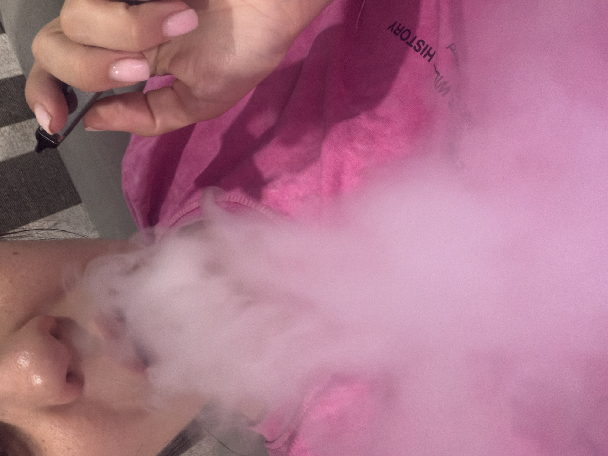 A close-up, low-angle shot shows a person exhaling a cloud of pink vapor from their mouth. The person's nose and lips are visible, along with a portion of their hand holding a vaping device. The person is wearing a bright pink t-shirt with black text printed on it, which is partially obscured by the vapor. The background is out of focus, appearing to be a couch with a striped pattern. The image appears to have been taken indoors. There is no indication of the city of Sochi, Russia.