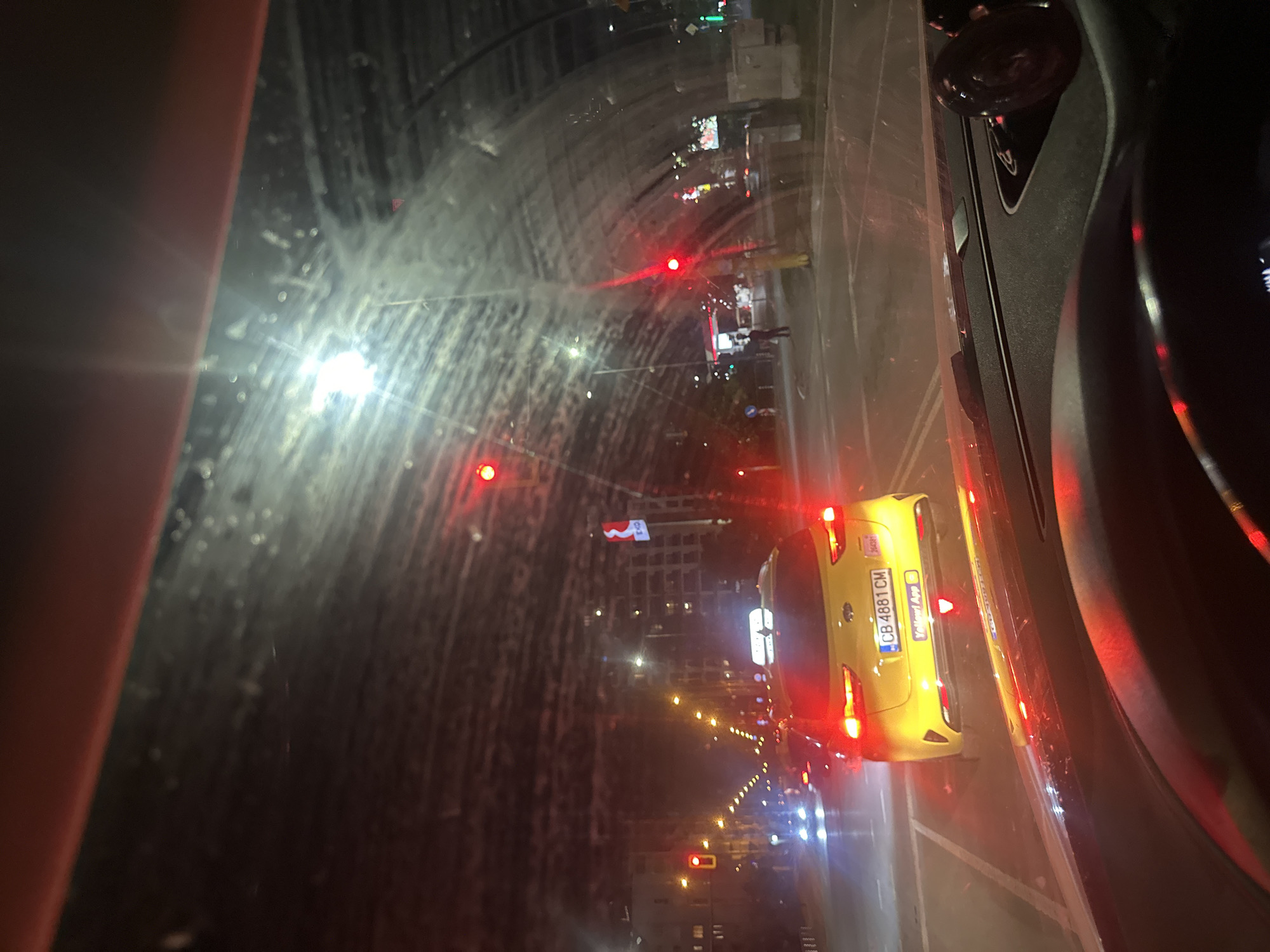 The image is taken from inside a vehicle at night, looking through the windshield. The windshield has water droplets and smudges on it, causing some glare and distortion. In the foreground, the dashboard and steering wheel of the car are visible. The main subject of the image is a yellow taxi with the license plate "CB 4881 CM" in the middle distance. The taxi has its brake lights illuminated and a light on its roof. The scene is a city street with multiple traffic lights, some of which are red, indicating traffic is stopped. Buildings and streetlights are visible in the background, suggesting an urban environment. The overall impression is that the car is stopped at a traffic light in Sofia, Bulgaria, at night.
