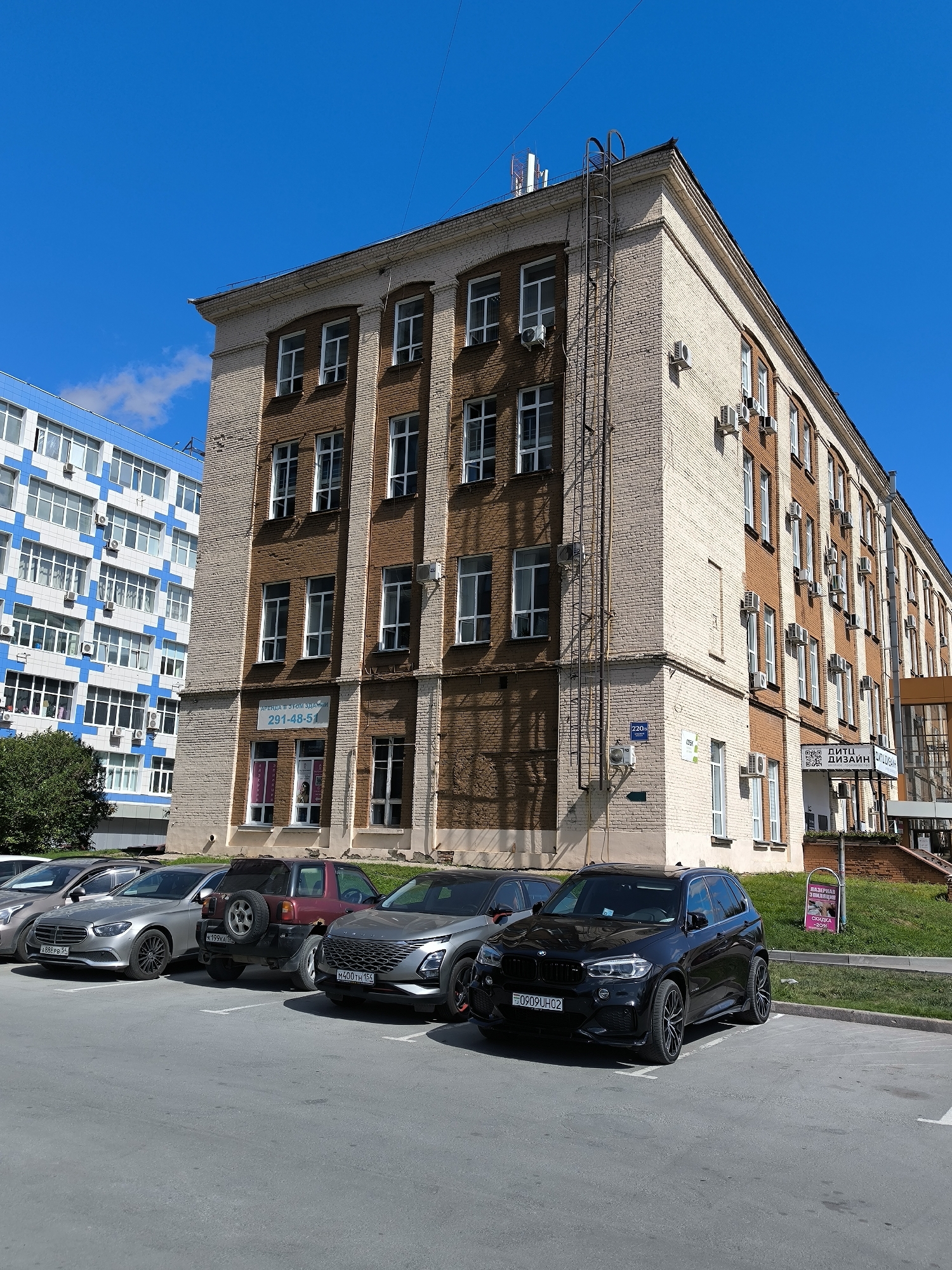 The image shows a side view of a multi-story brick building with white trim and numerous windows. The building is located in Novosibirsk, Russia, as indicated by the presence of a sign that reads "ДЕТИ ДИЗАЙН" and the license plates of the cars, which are consistent with Russian plates. The building is situated next to a modern, blue and white building with many windows. In front of the brick building, there is a parking lot with several cars parked, including a black BMW SUV, a silver sedan, and a red SUV. A ladder is attached to the side of the brick building, and there are several air conditioning units mounted on the exterior walls. The sky is clear and blue, and there is a small cloud visible in the upper left corner of the image. The overall scene depicts an urban environment on a sunny day.