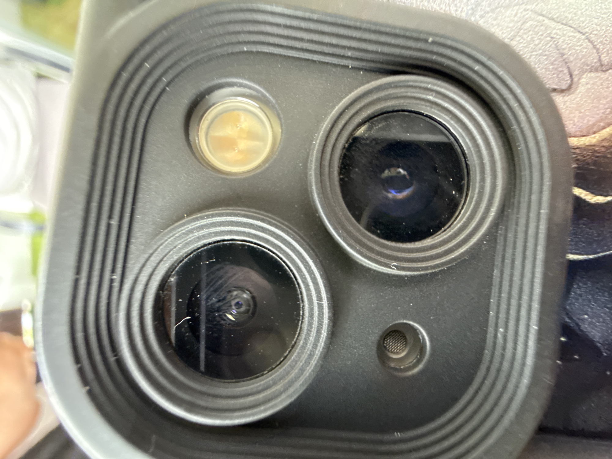 This close-up image, captured in Garmisch-Partenkirchen, Germany, displays a dark grey or black rectangular camera module, likely belonging to a smartphone or similar electronic device. The module's surface is textured and surrounded by concentric raised lines. Within this module, two primary circular camera lenses are visible: one larger lens in the upper-left quadrant and a slightly smaller lens in the lower-right quadrant. Both lenses exhibit reflections and an accumulation of fine dust particles and several white fibers on their surfaces.
To the upper right of the larger lens, a small, circular flash unit is present, emitting a yellowish light and revealing two distinct internal LED elements. Below the larger lens and to the lower left, a smaller circular recess contains a fine mesh grille, indicative of a microphone or sensor port.
The background elements are heavily blurred due to shallow depth of field. The upper portion of the background shows indistinct shapes and colors, including a warm skin tone, white, and green. The lower background features blurred patterns of light beige/brown and darker blue/purple, suggesting a soft, patterned surface.