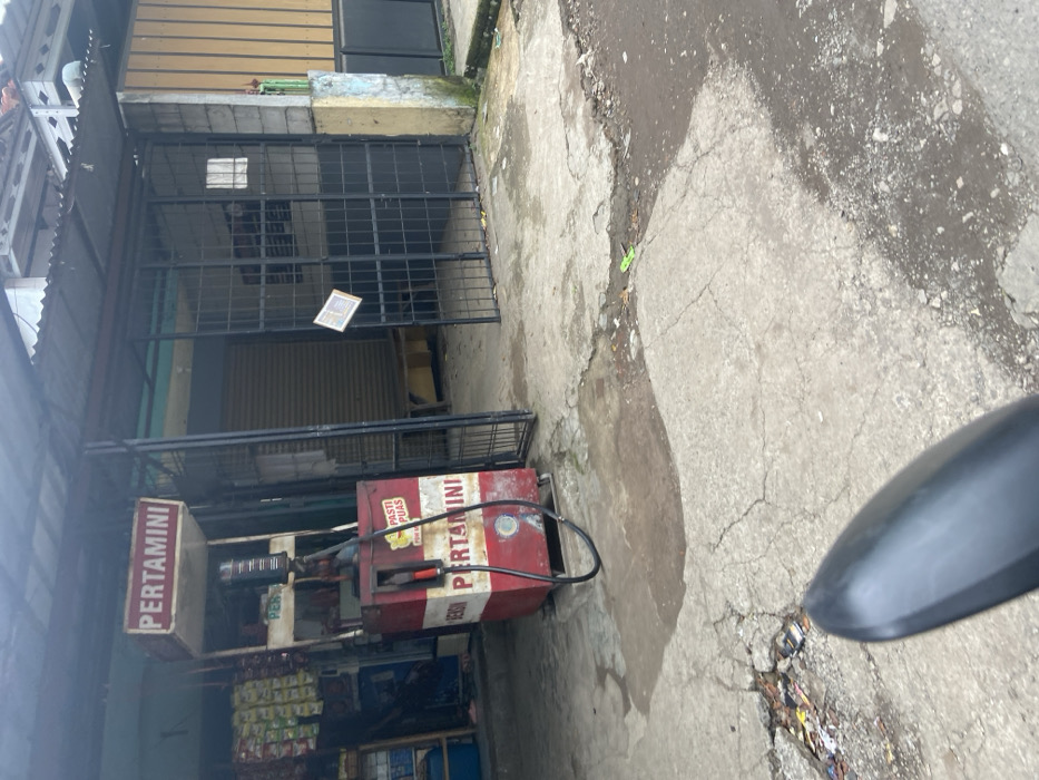 The image shows a gas station in Baleendah, Indonesia, located at the coordinates -7.021 latitude and 107.677 longitude. The gas station is a small, standalone structure with a red and white sign that says "Pertamini" in Indonesian. A single gas pump is visible, and the station has a small, enclosed area with a gate. The pump is red and white with a sign that reads "Pasti Puas" (Guaranteed Satisfaction). The gas station is located on a narrow, paved street. There is a black motorcycle in the foreground, with its mirror visible in the bottom right corner of the image. The road surface is cracked and uneven. The area around the station is dusty and appears to be in an urban area. There are no people visible in the image.