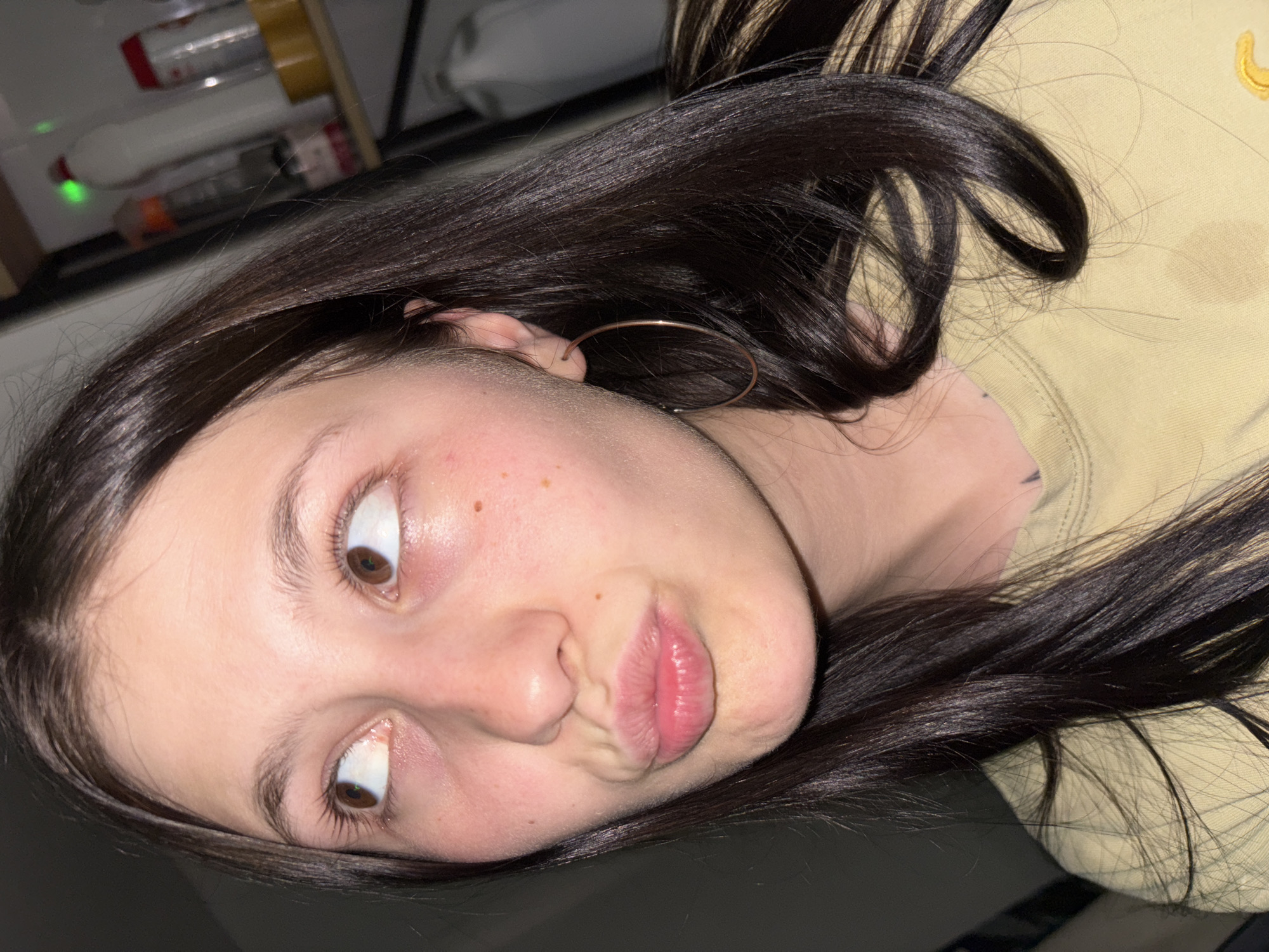 A close-up, slightly angled photograph shows a young woman with dark brown hair and light skin making a pouting expression. She has brown eyes that are looking upwards and slightly to her left. She is wearing a light-colored shirt and a hoop earring. The background is blurred and includes glimpses of objects on a shelf. The location in the city of Фоки, Russia cannot be confirmed.