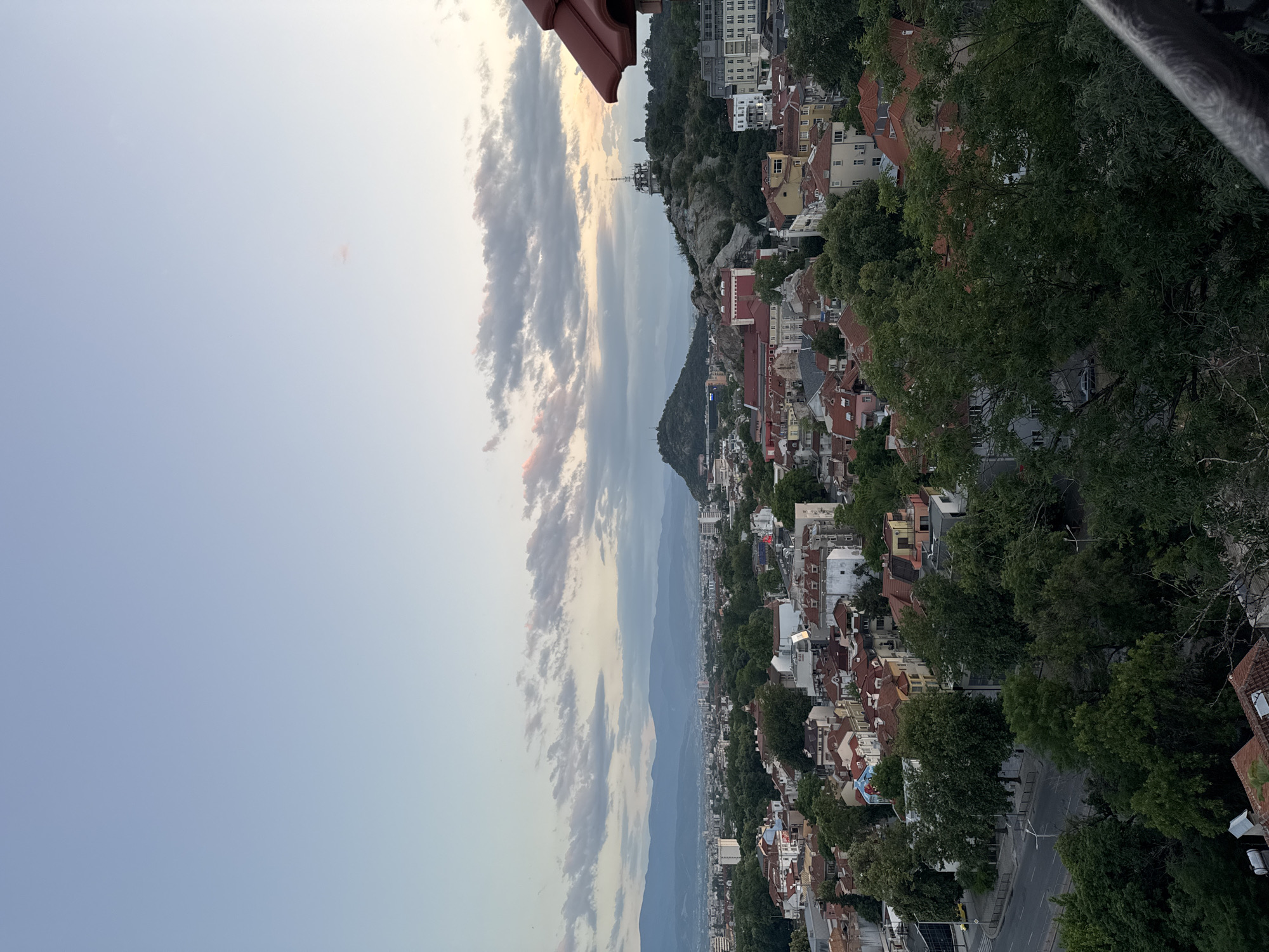 This is an elevated, panoramic view of the city of Plovdiv, Bulgaria, captured during the late afternoon or early evening twilight. The scene is bisected horizontally by the cityscape, with the upper two-thirds dominated by the sky and the lower third by the urban environment and foreground elements.
The sky exhibits a gradient from light blue at the zenith to a paler, more muted blue-grey near the horizon. Scattered stratocumulus clouds are present across the mid-sky, displaying subtle pink and orange coloration on their undersides, indicative of indirect sunlight from a setting or recently set sun. A distinct, hazy blue mountain range forms the distant horizon line.
The urban landscape in the midground features a dense concentration of buildings with varying architectural styles, predominantly characterized by numerous structures with terracotta-tiled roofs. Extensive green tree canopy, indicative of deciduous foliage, is interspersed throughout the city, forming parks and lining streets.
Two prominent hills, or 'tepes,' are discernible within the city:
1. On the right side of the frame, Bunardzhika Hill (Hill of the Liberators) rises, topped by a large, multi-level communication tower and the silhouette of a monumental statue (Alyosha Monument). Its slopes show rocky formations and dense tree cover.
2. Further to the left, a second, less prominent, tree-covered hill (likely Sahat Tepe or Dzhendem Tepe) is visible above the city's general elevation.
In the lower-left quadrant, a two-lane road is visible, partially obscured by mature trees. A single yellow vehicle is identifiable on this road. The immediate foreground on the lower right reveals a dark, textured metal balustrade or railing, suggesting the observation point is from an elevated structure or another tepe, such as Nebet Tepe, looking west-northwest across the city. No human figures are discernible in the image.