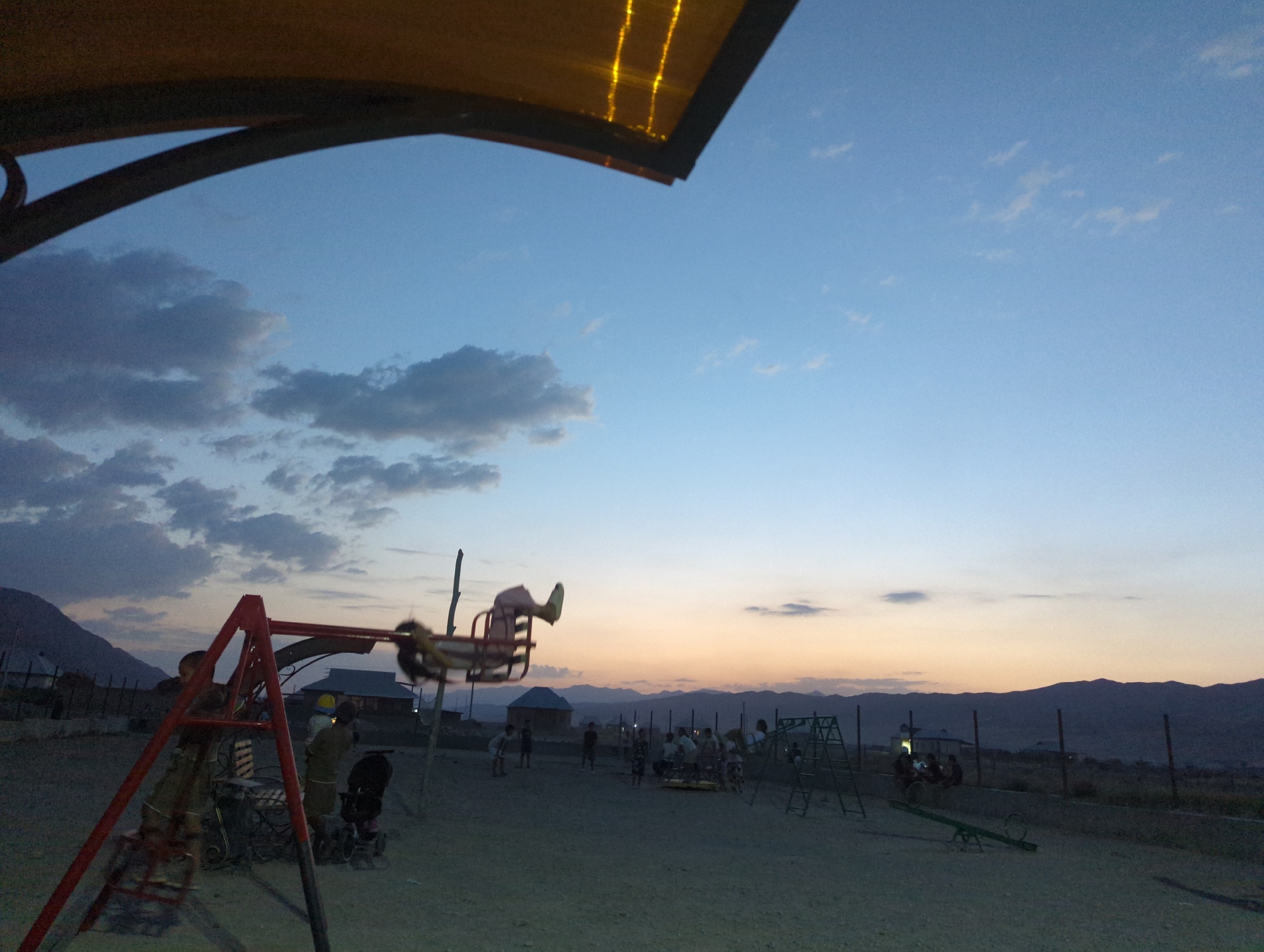 This image captures a playground scene at dusk in the village of Üç-Döbö, Kyrgyzstan. The sky is a gradient of pale blue to orange as the sun sets behind a range of dark mountains in the distance. In the foreground, a red metal swing set stands, with one swing occupied by a blurry figure in motion. To the right of the swing set, a group of children are gathered around a merry-go-round. Further to the right, a green metal ladder structure for climbing is visible. The ground is a dusty, sandy texture. Several simple houses and fences are scattered throughout the background, interspersed with trees and sparse vegetation. The overall atmosphere is one of casual, late-day recreation in a rural setting.