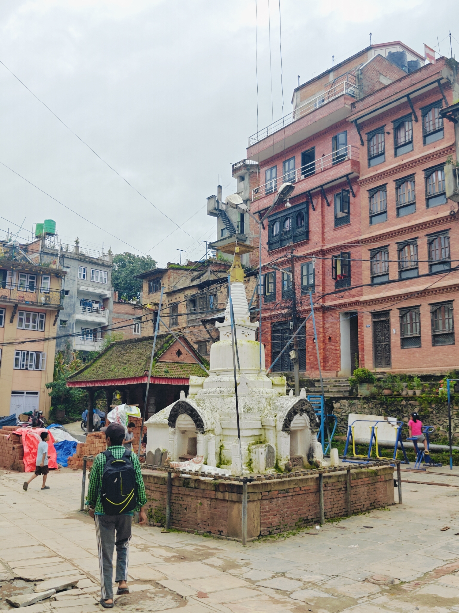 In Kirtipur, Nepal, a man wearing a green checkered shirt and a backpack walks away from the camera on a paved stone courtyard. To his left, a white stupa with brick detailing stands in the foreground. Behind the stupa, a red brick building with multiple stories and traditional windows is visible. To the right of the stupa, a playground set with yellow and blue bars is present, with a young person in a pink shirt using some of the equipment. In the background, more buildings of varying architectural styles and heights, along with trees, complete the urban landscape under an overcast sky. The scene appears to be a public space within the city.
