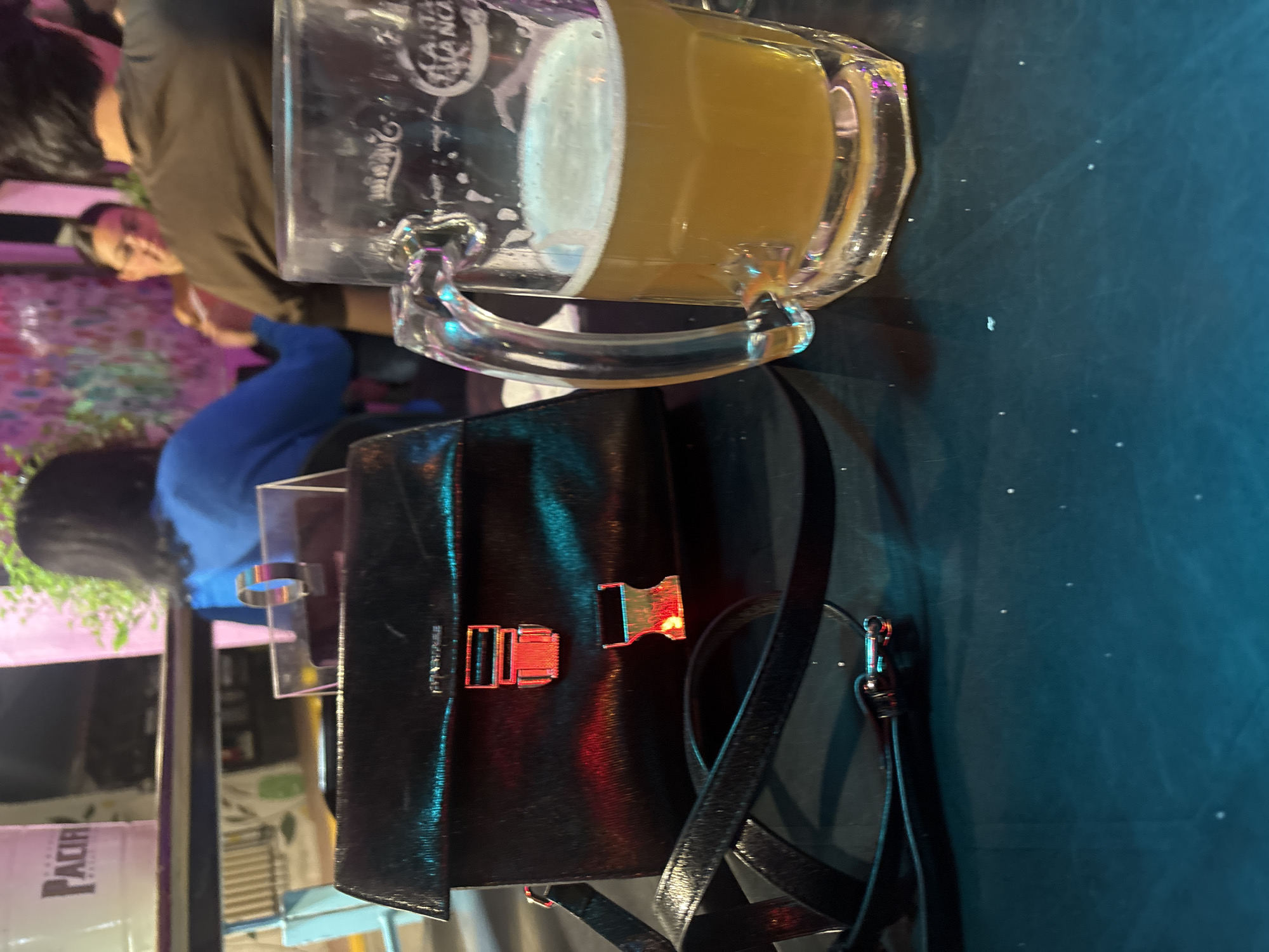 A still life scene captured in the city of San Andrés Cholula, Mexico, features a glass mug filled with beer, next to a black purse. The table is dark with scattered bright dots, and glimpses of people are seen in the background. The lighting appears dim, possibly indicating an indoor setting like a bar.