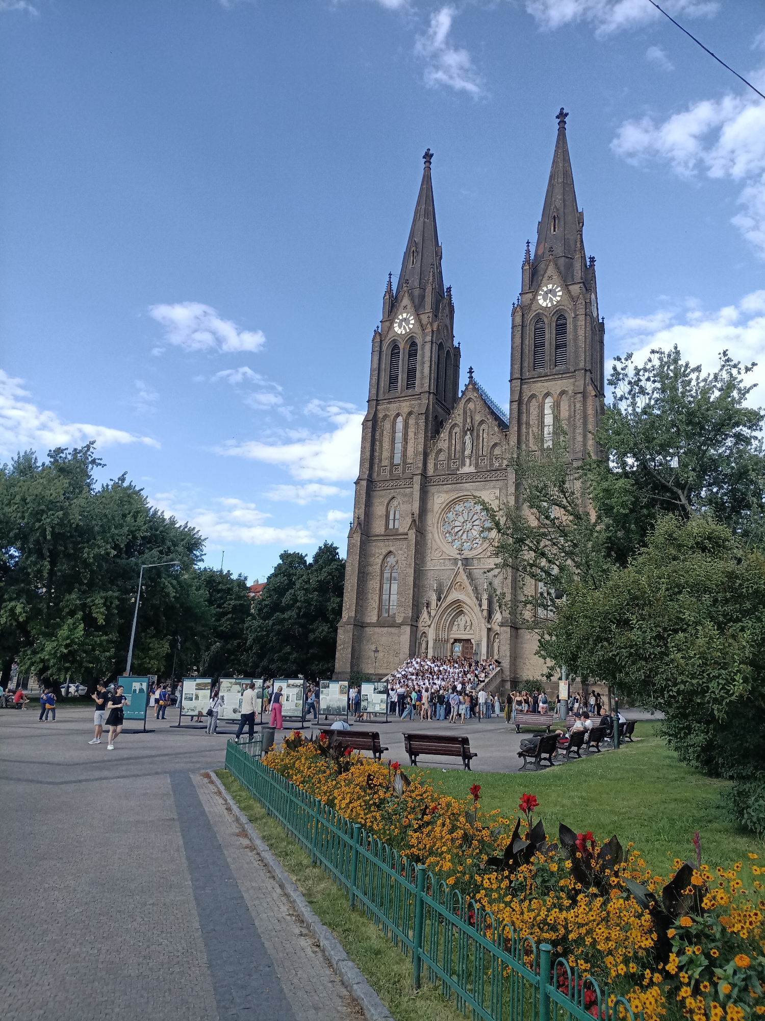 The image shows the Basilica of Saint Peter and Saint Paul in Vyšehrad, Prague, Czechia. The basilica features twin spires, a large rose window above the entrance, and intricate gothic architecture. In front of the basilica, a large group of people are gathered on the steps and in the plaza below. A park area with benches and flower beds borders the plaza. The sky is blue with scattered white clouds. Various trees surround the basilica and park area.