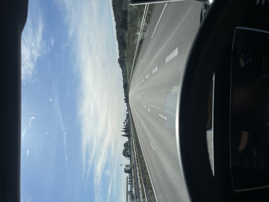 The image captures a driver's perspective from within a vehicle traveling on a multi-lane highway in Tavel, France. In the immediate foreground, the top section of a black steering wheel is visible, with a partially obscured digital dashboard screen below it. The view through the windshield shows a multi-lane asphalt road extending straight towards the horizon under a bright blue sky with scattered white clouds. White dashed lane markings are present on the asphalt surface. To the left of the highway, a metal crash barrier runs parallel to the road, beyond which a gas station facility is visible, comprising multiple fuel pumps under a canopy and an adjacent building. A line of tall, slender trees borders this area. To the right of the highway, another metal crash barrier separates the road from a grassy embankment sloping upwards, populated with scattered trees. No other vehicles or individuals are explicitly visible within the frame. The scene is illuminated by clear daylight.