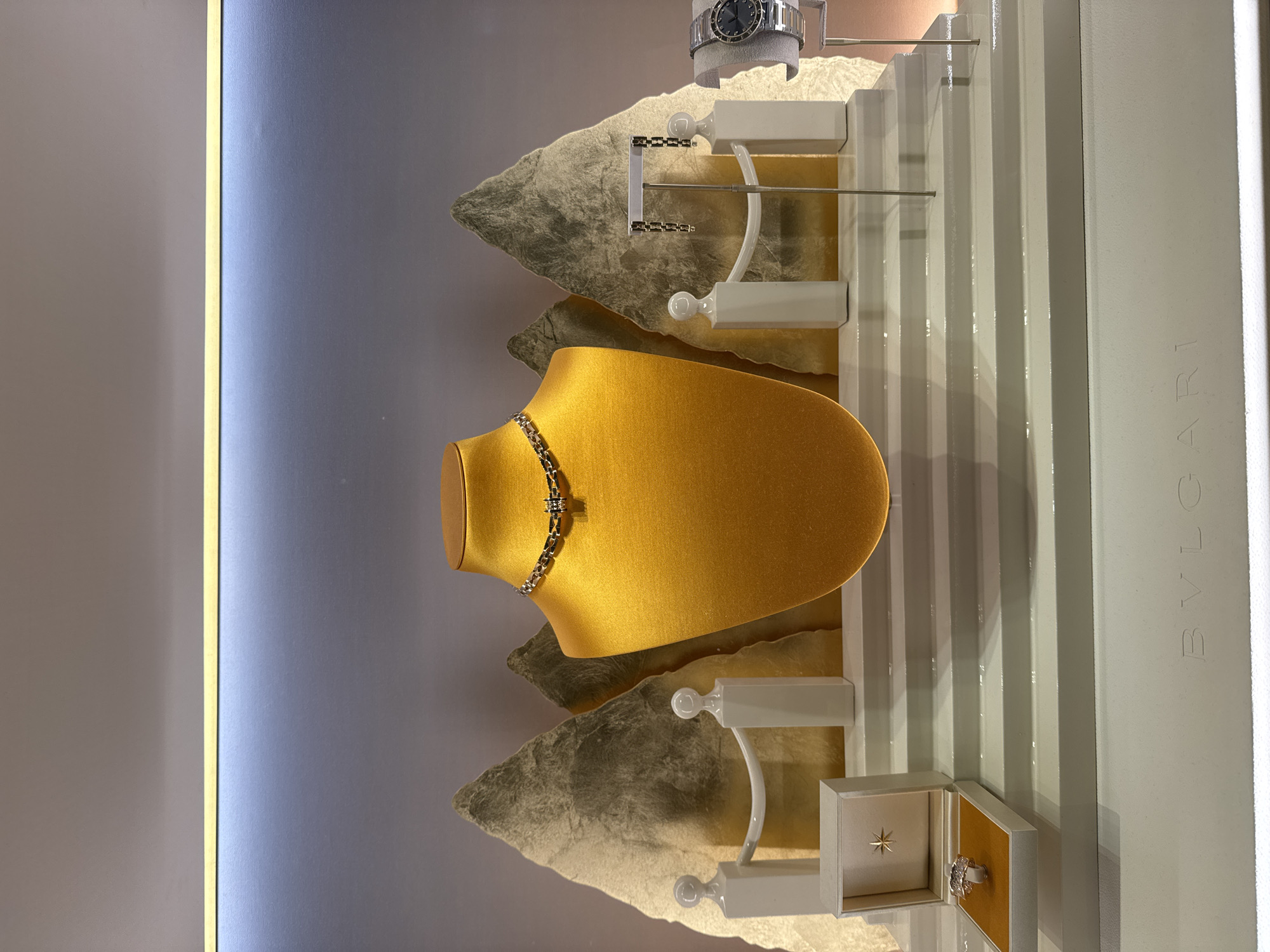 The image shows a high-end jewelry display inside a store. The display features a gold-colored mannequin bust wearing a silver necklace. To the left, a ring is presented in an open white box. To the right, a pair of earrings hangs from a stand, and a watch is displayed on a gray cushion. The backdrop consists of two abstract, textured golden shapes resembling mountains, with white decorative posts and railings. The display is set on multiple white tiered platforms. At the bottom of the image, the word "BVLGARI" is subtly embossed on the display's base. The lighting is soft and highlights the jewelry. The scene is set in a retail environment, likely a jewelry store, potentially located in the city of Nong Prue Subdistrict, Thailand, given the request. There are no people visible in the image.