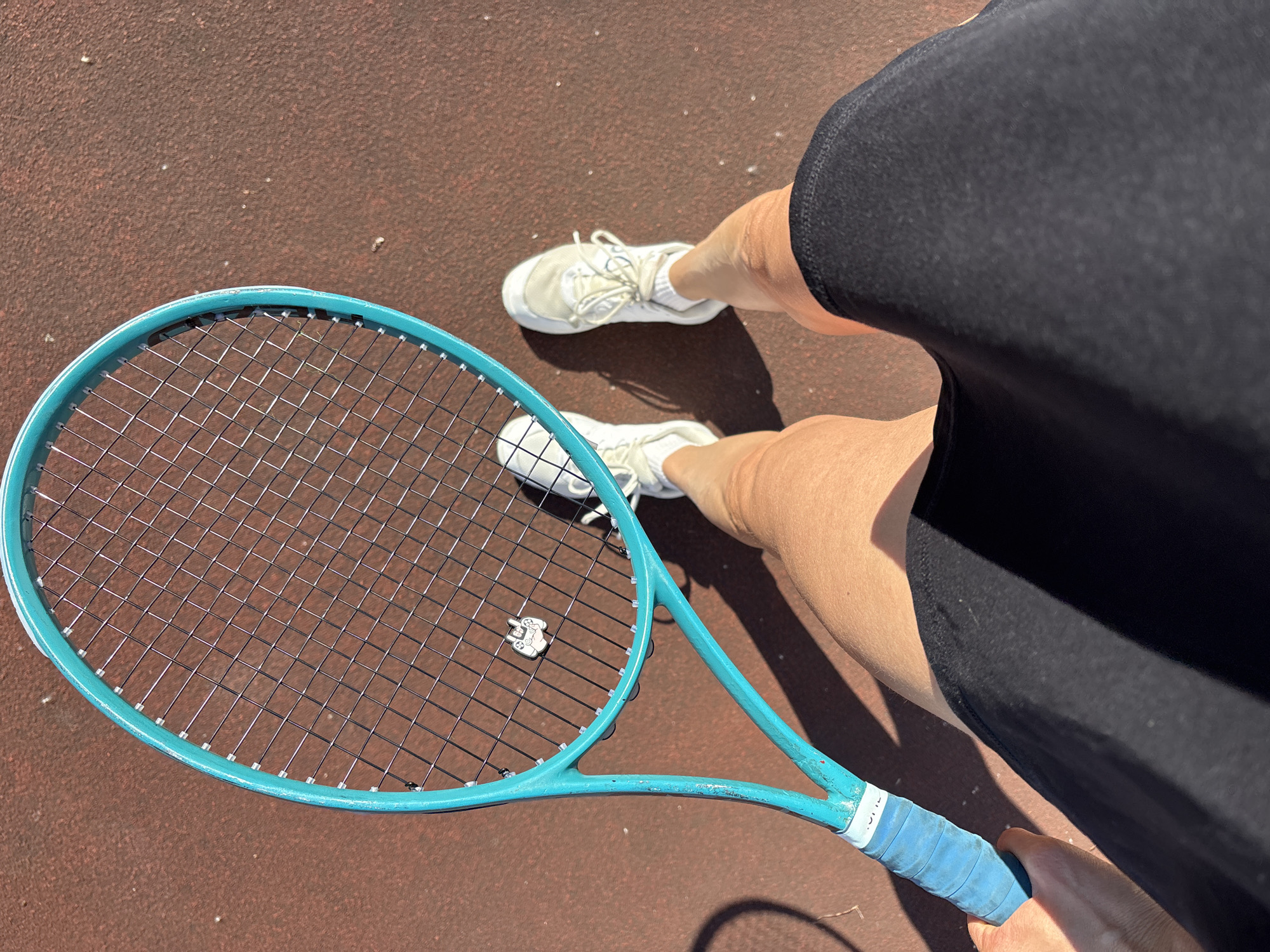 A first-person perspective shot shows a person on a tennis court holding a teal tennis racket. The racket has a blue grip and a small white charm attached to the strings. The court surface is a reddish-brown color. The person is wearing white sneakers and a black skirt. Their legs are visible, along with a shadow cast by sunlight. The image does not provide any information that would allow for identification of the location within Chaikovsky, Russia.