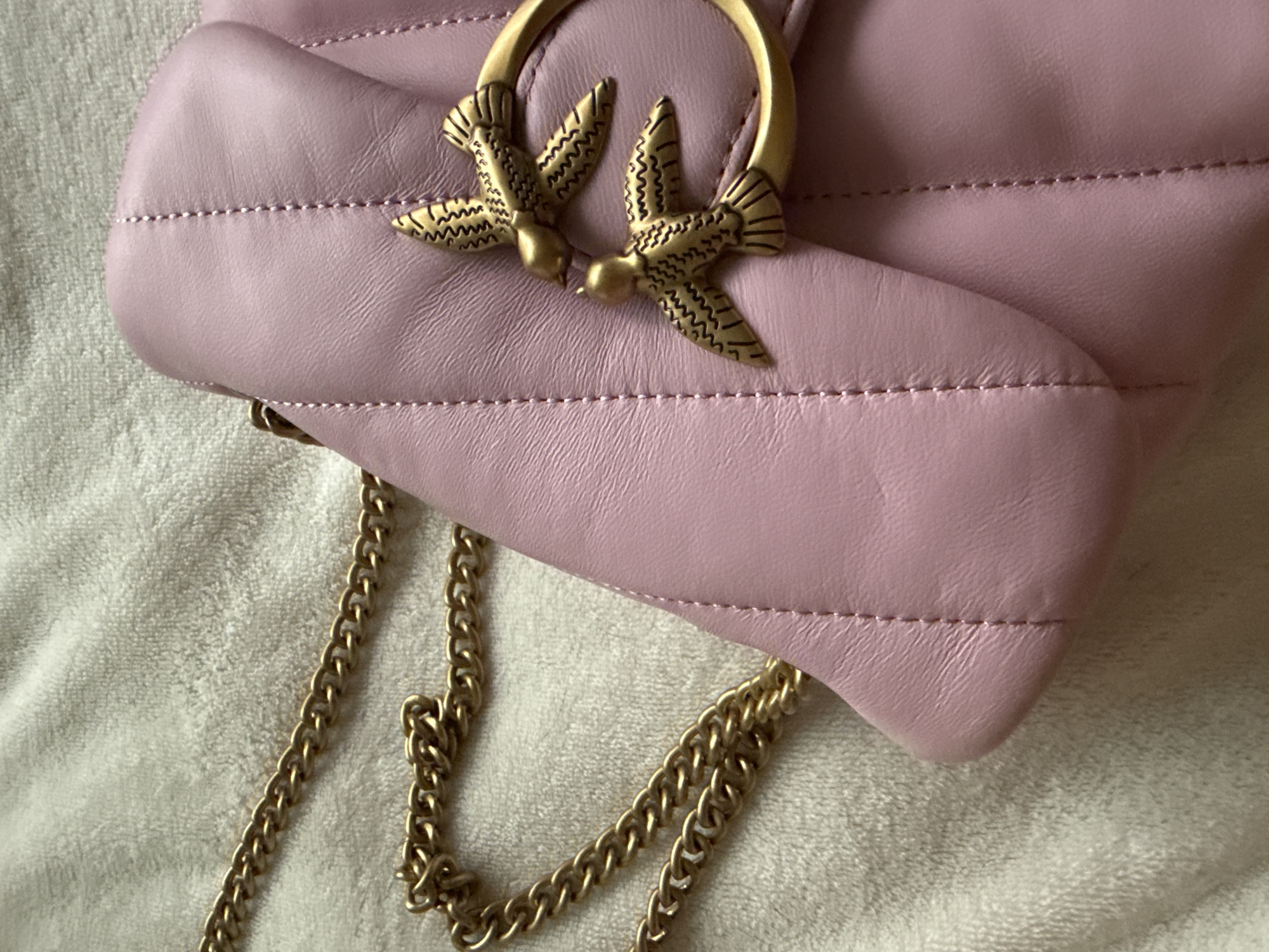 The image shows a close-up, high-angle view of a pink, quilted leather handbag with a gold chain strap. The bag is adorned with two gold bird-shaped clasps. The texture of the bag and the chain are clearly visible. The background is a cream-colored, textured fabric. The image is taken indoors and does not contain any elements that would indicate a specific location in Bad Sachsa, Germany.